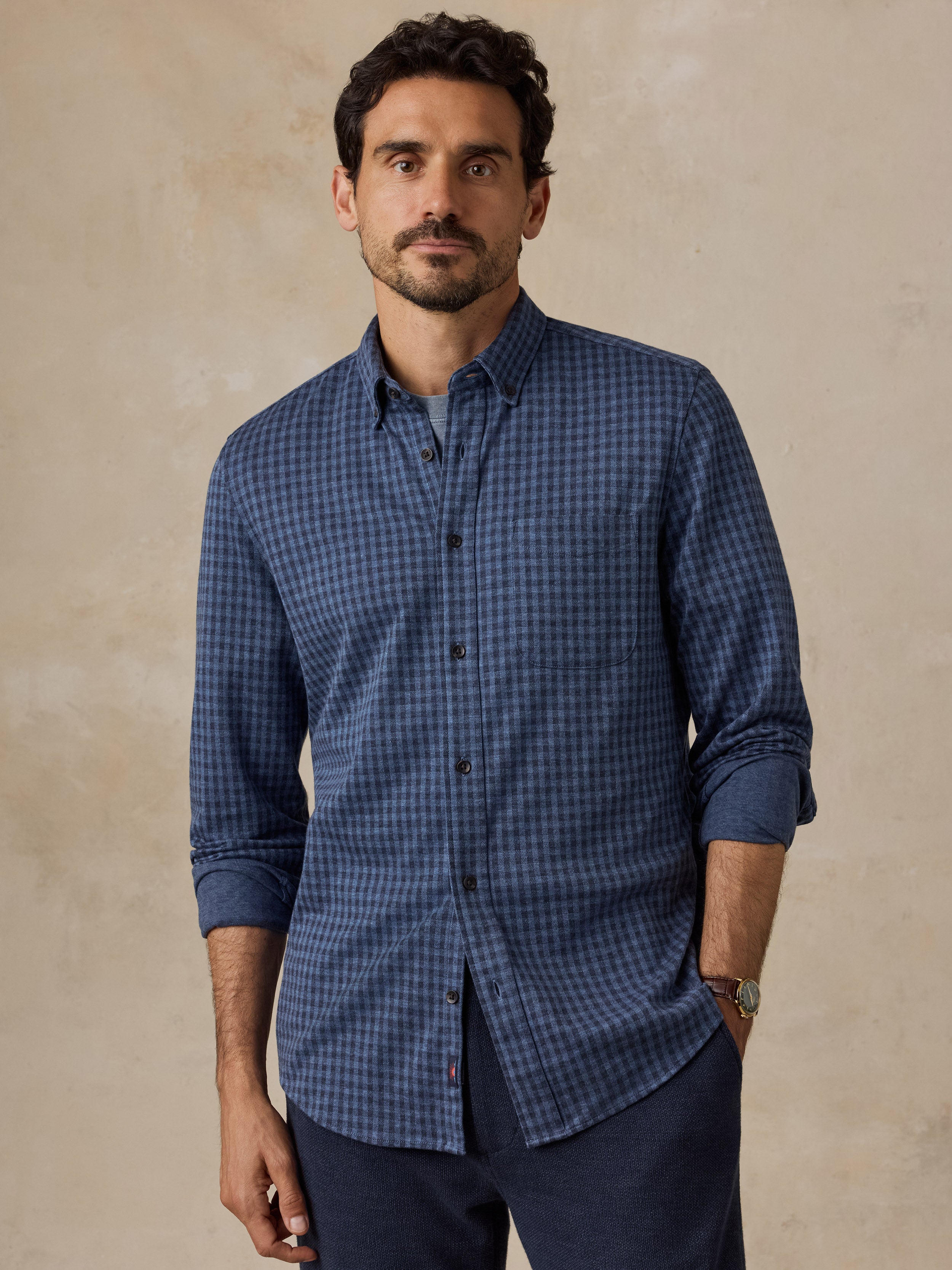 A man wearing a blue and navy checkered long-sleeve button-down shirt with dark navy trousers, standing against a textured beige background.