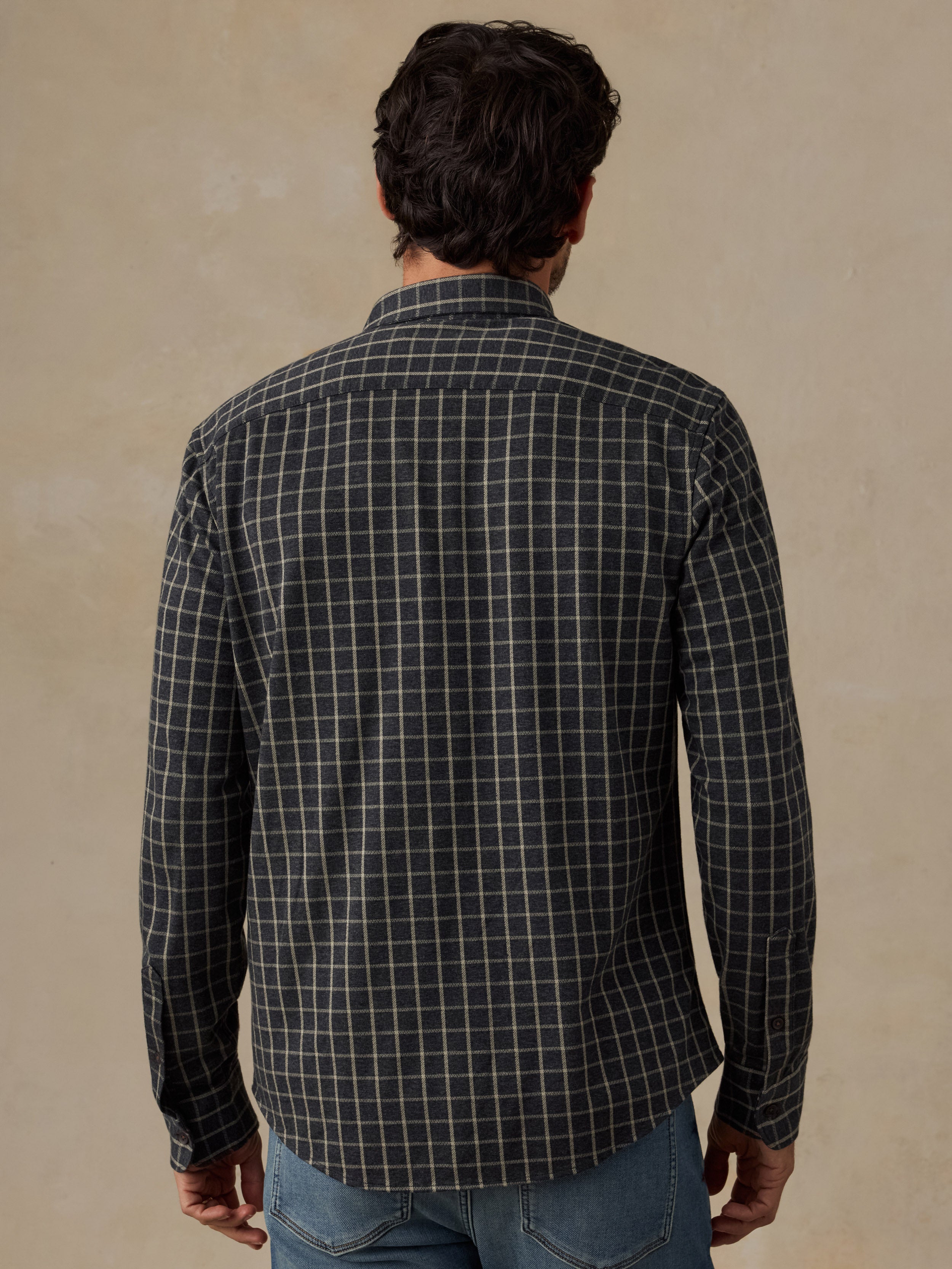 A man viewed from behind wearing a dark charcoal gray long-sleeve button-down shirt with a white windowpane plaid pattern, paired with blue jeans, against a textured beige background.