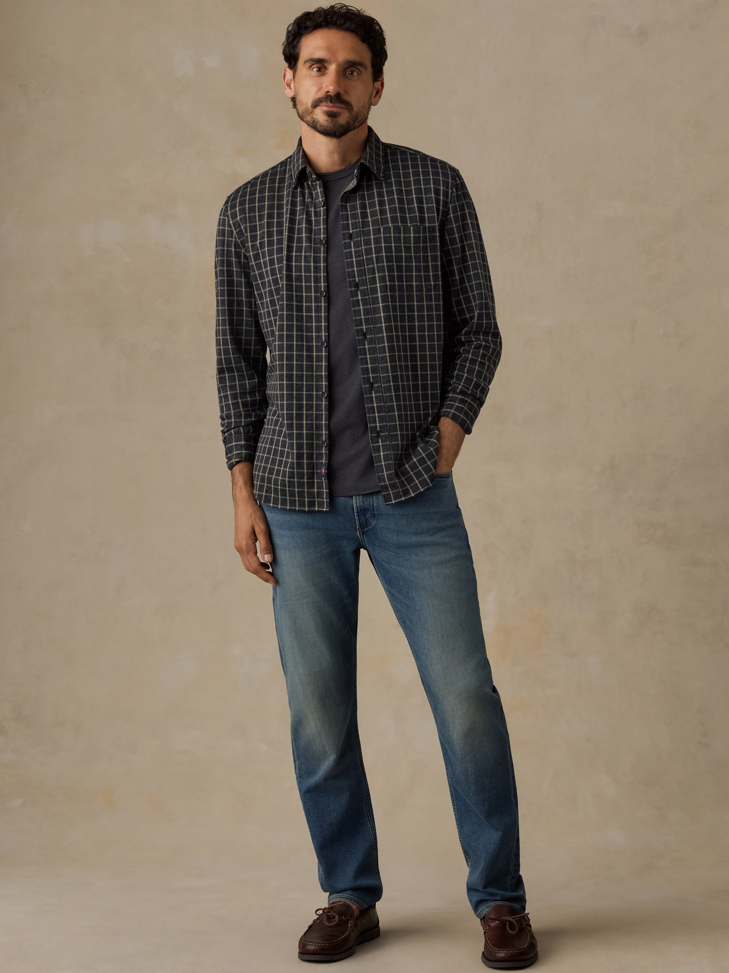 A man modeling a dark gray and black plaid button-down shirt over a dark t-shirt, paired with blue jeans and brown leather shoes, standing against a textured beige studio backdrop.