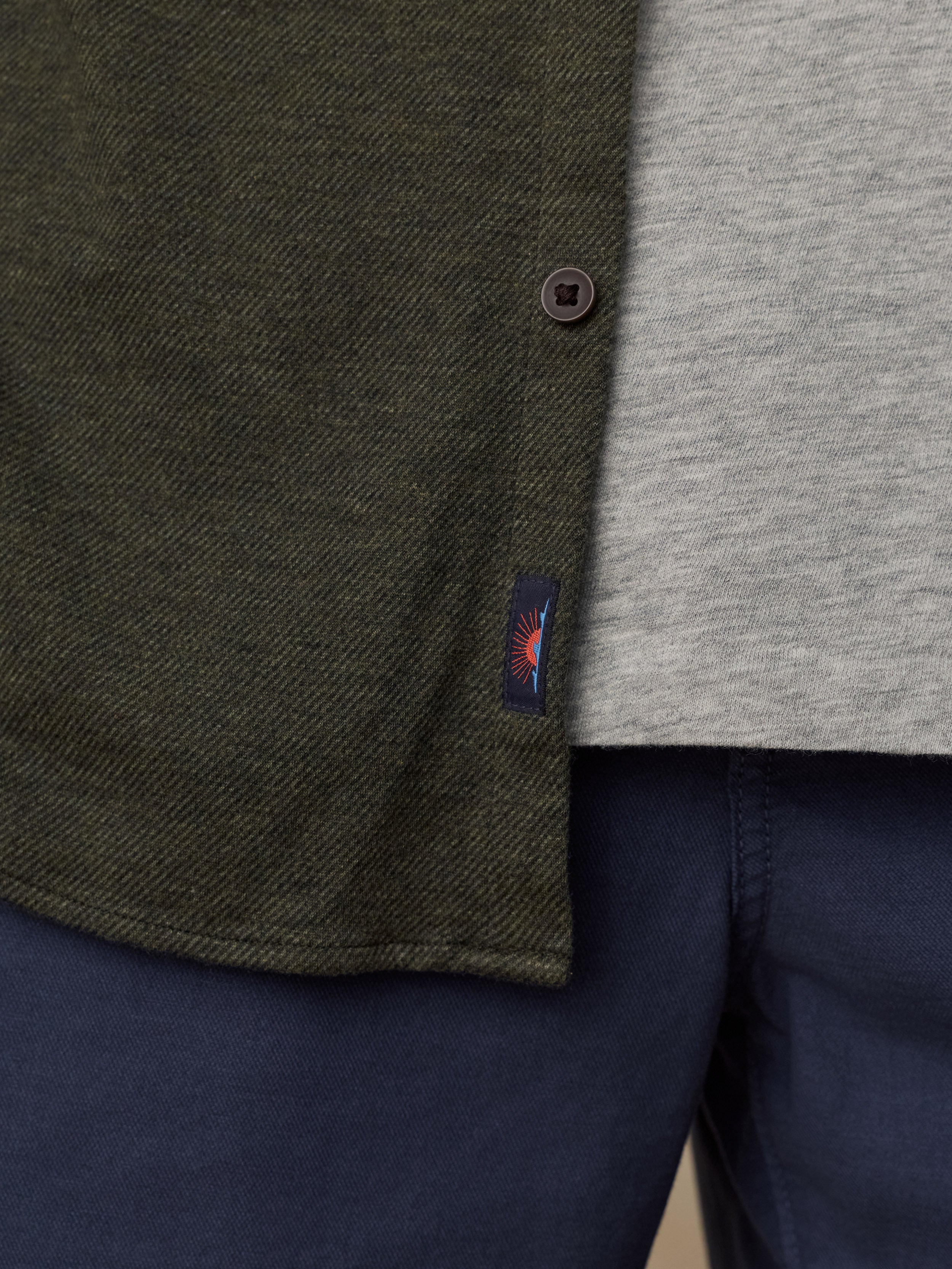 Close-up of a man wearing an olive green textured overshirt over a light gray heathered t-shirt and navy blue trousers, with a small Faherty brand tag visible on the overshirt.