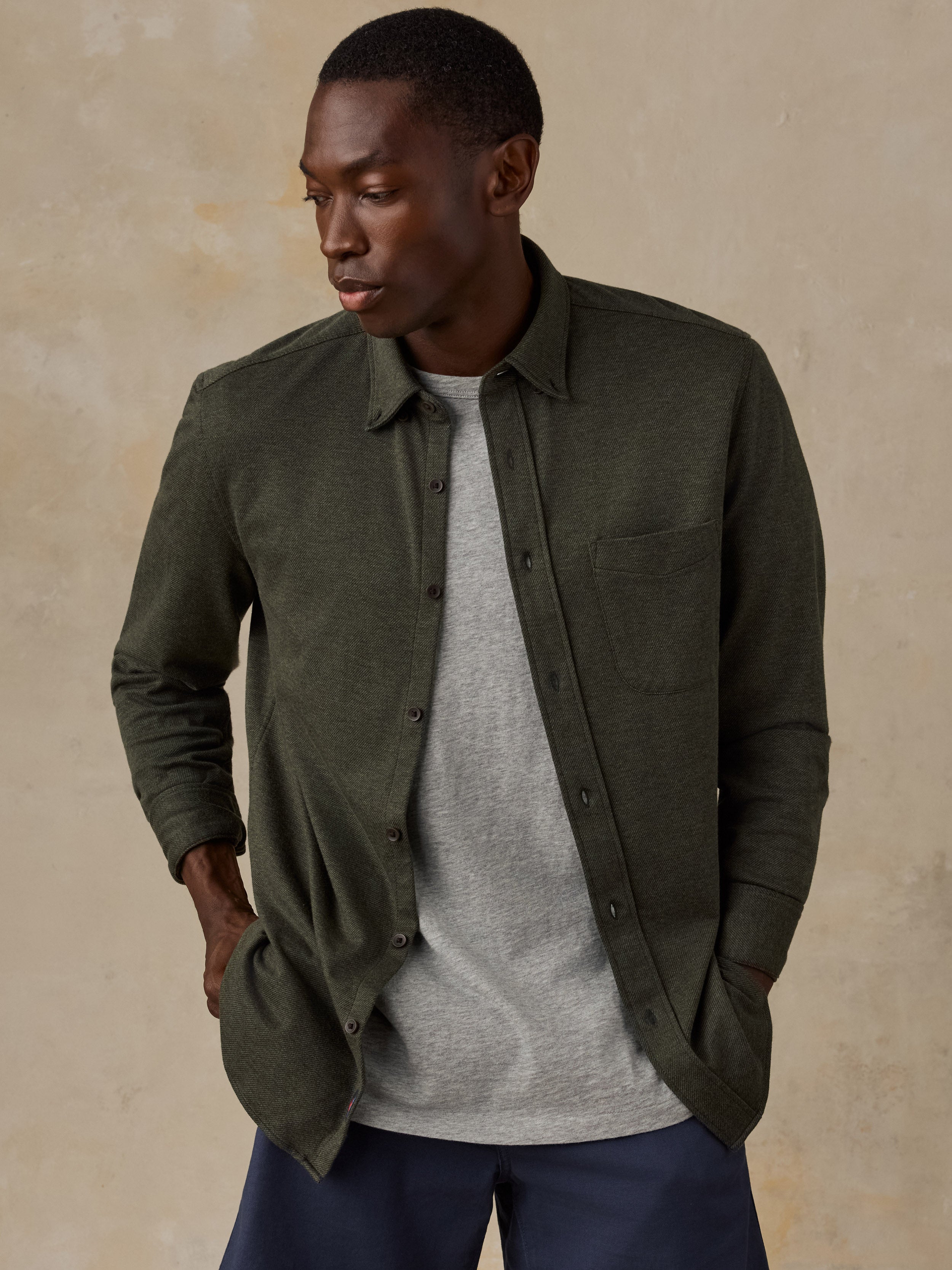 A man wearing an olive green long-sleeve button-up shirt over a light gray t-shirt and navy blue pants, standing against a textured beige background.