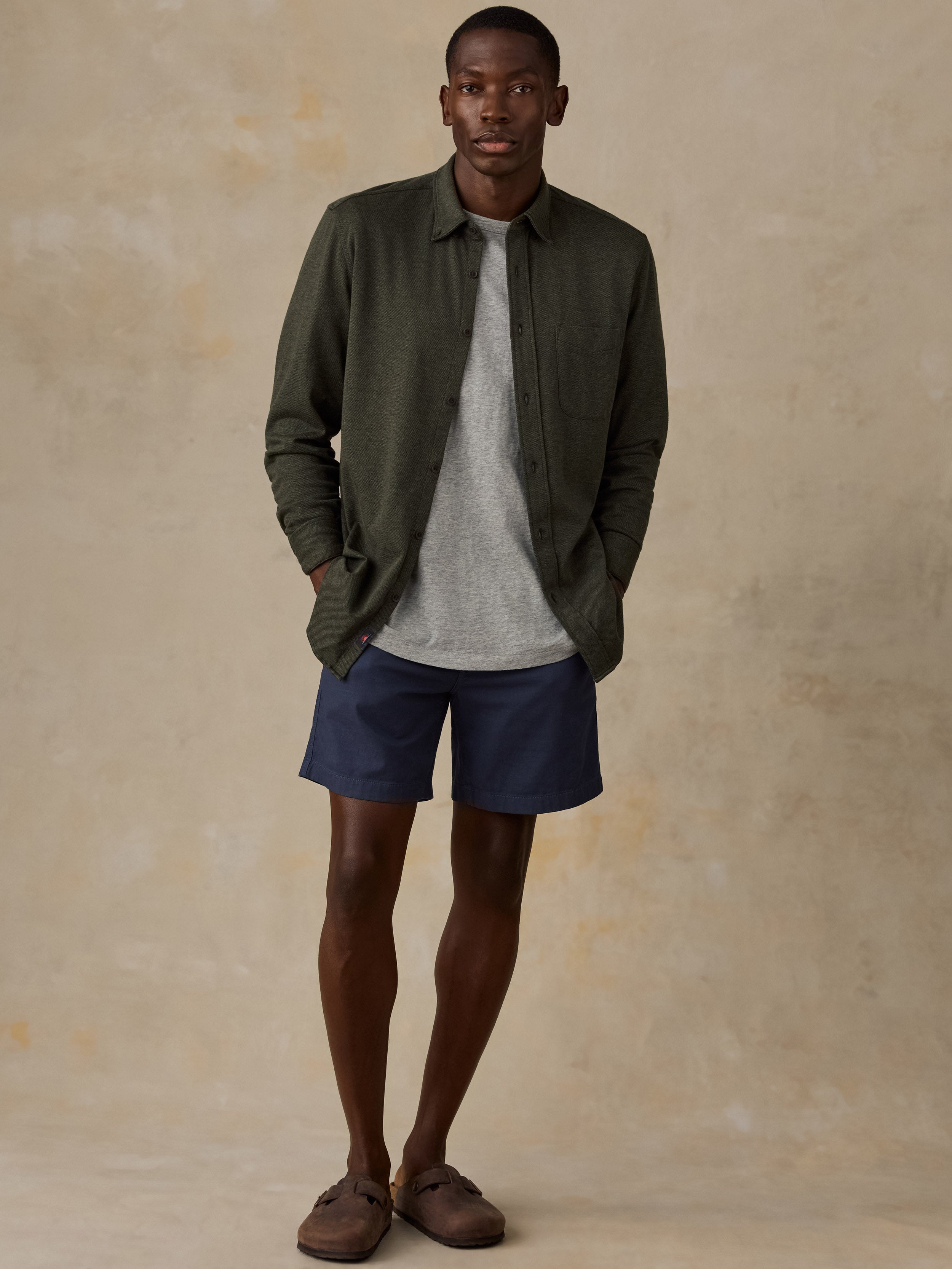 A man stands against a textured beige studio backdrop wearing an olive green button-up shirt over a light gray crewneck sweater, navy blue shorts, and brown slip-on shoes.