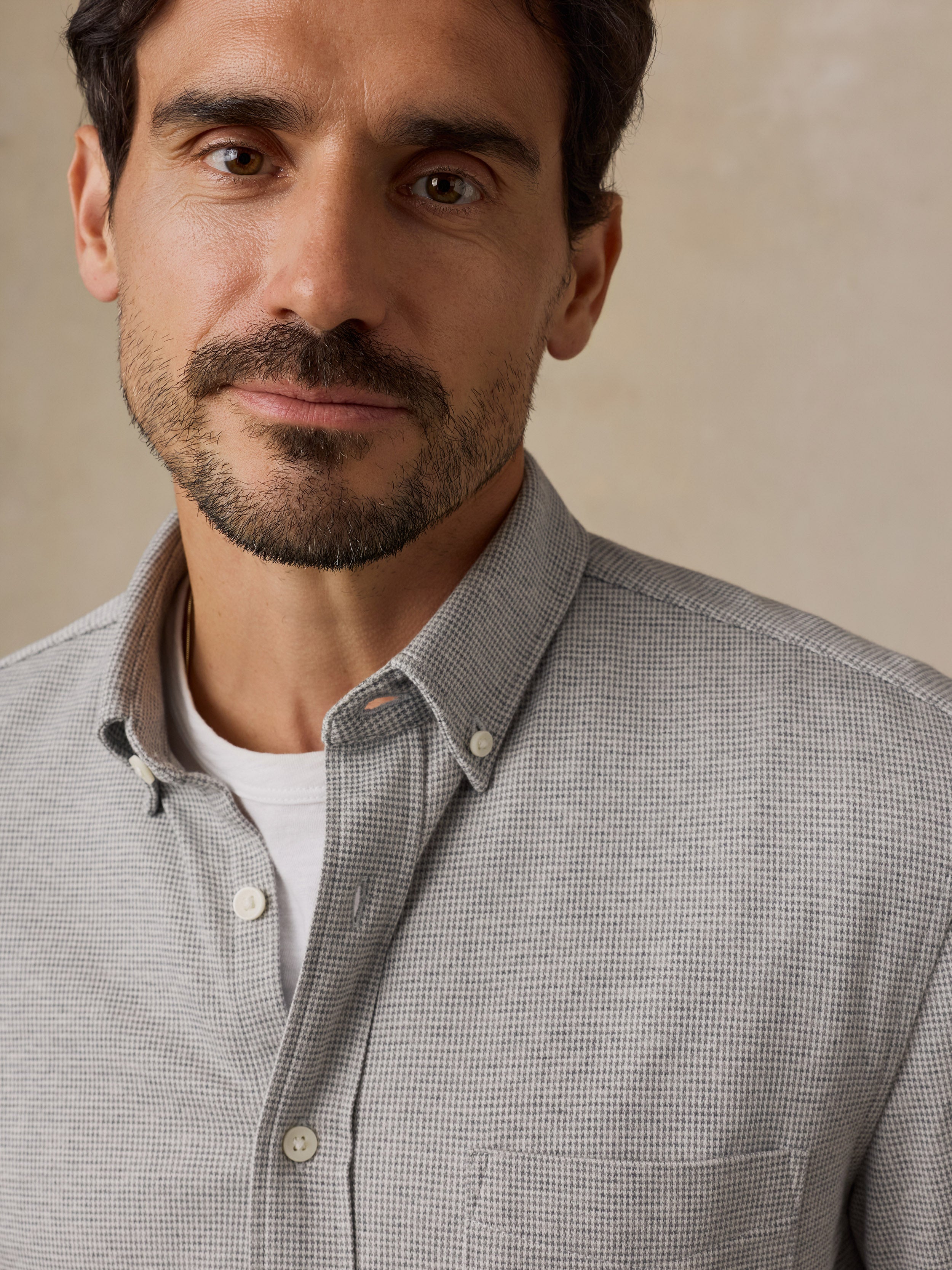 A close-up of a man wearing a light gray and white micro-check patterned button-down shirt over a white t-shirt, set against a neutral beige background.