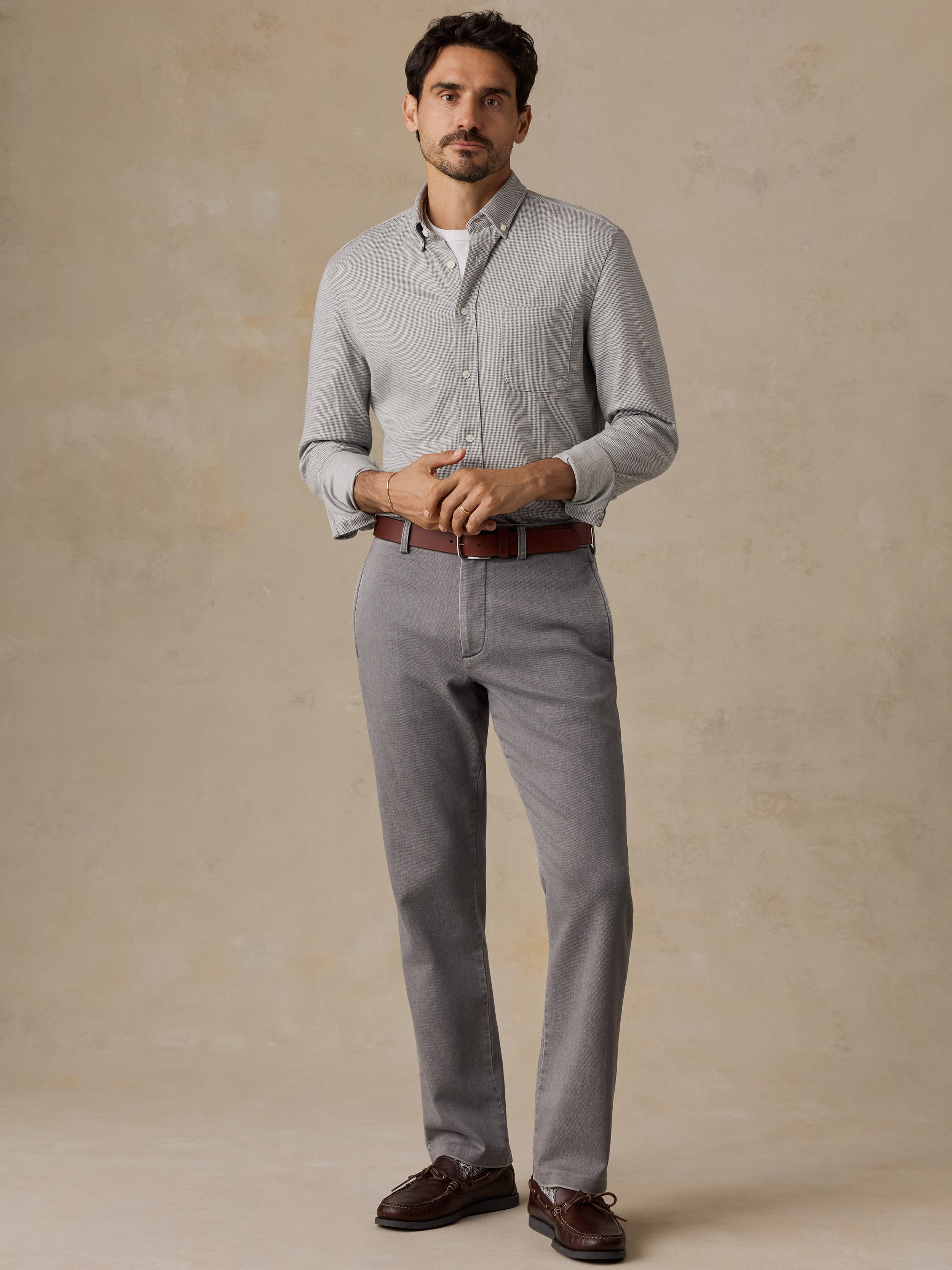 A man modeling a light gray textured knit cardigan over a white collared shirt, paired with gray trousers, a brown belt, and brown leather shoes, standing against a textured beige studio backdrop.