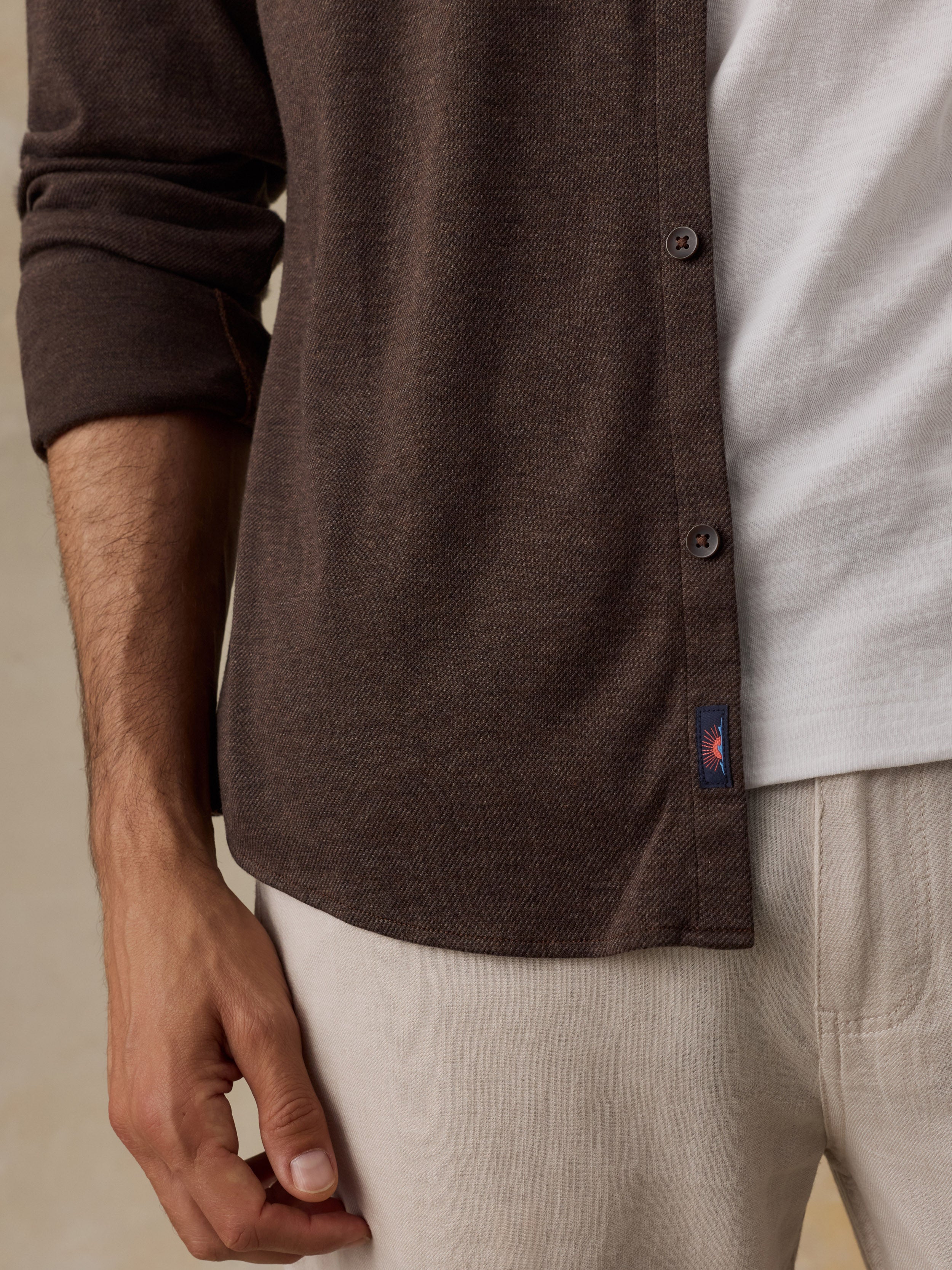 Close-up of a man wearing a dark brown long-sleeve button-up shirt over a white t-shirt and light beige trousers, showing the fabric texture and a small Faherty brand tag near the hem.