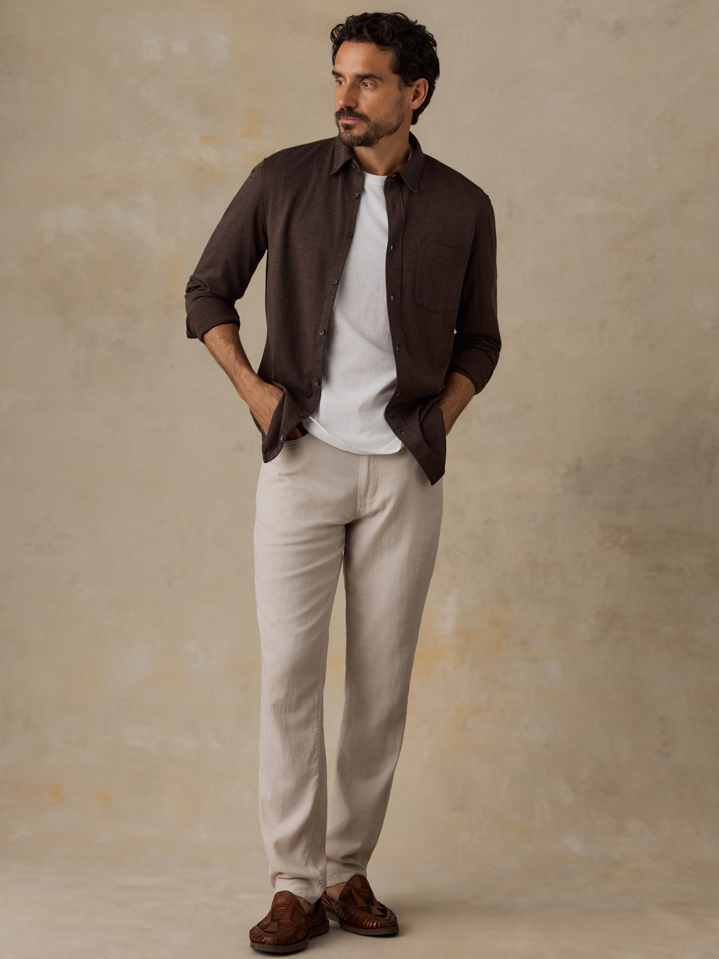 A man stands against a textured beige studio backdrop wearing a dark brown button-up shirt over a white T-shirt, light beige trousers, and brown loafers.