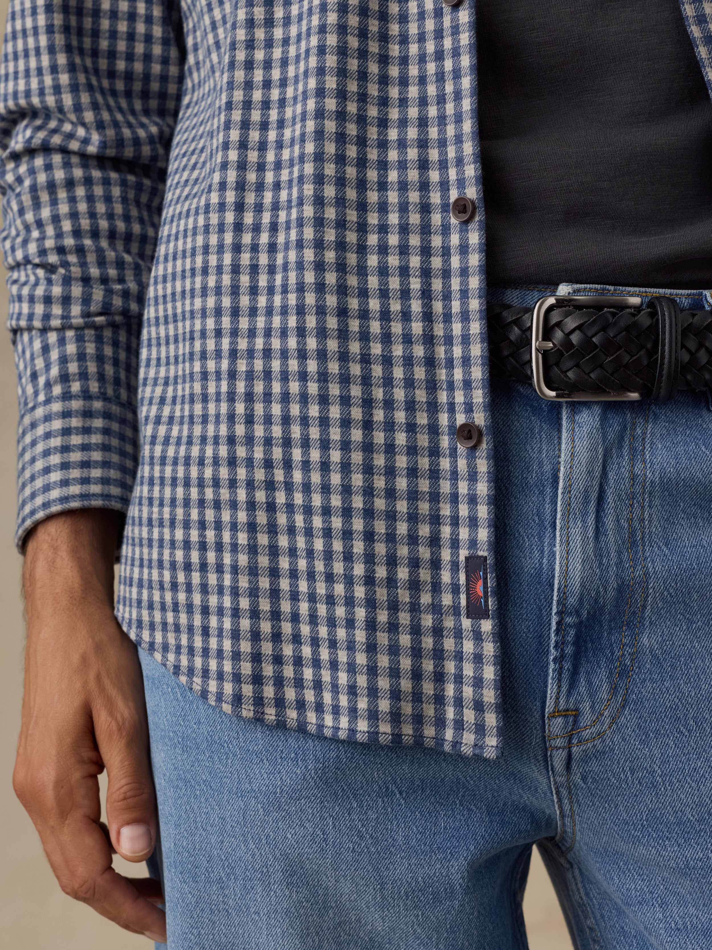 Close-up of a person wearing a blue and white gingham overshirt over a black shirt, paired with blue jeans and a black braided belt.