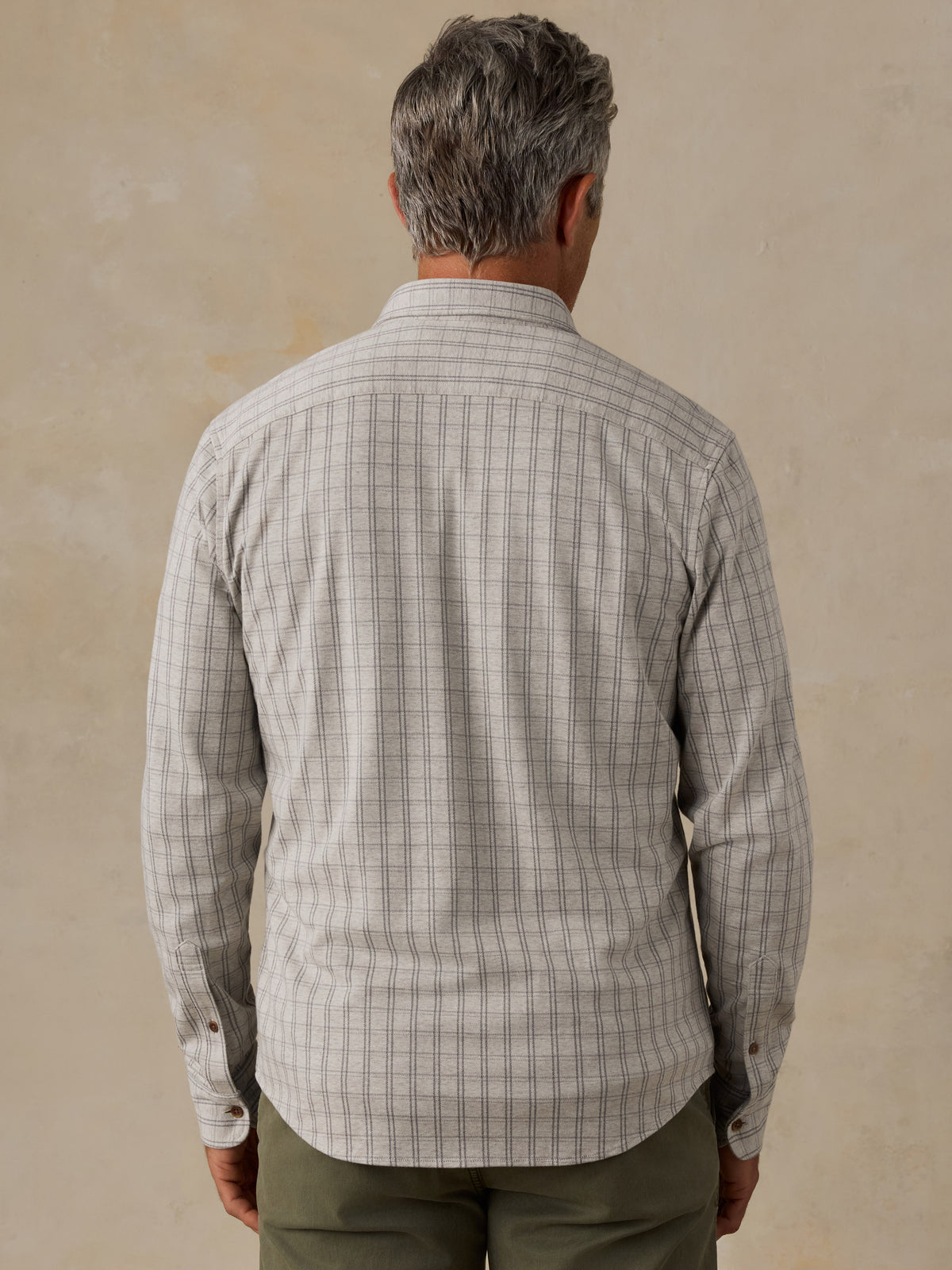 A man viewed from behind wearing a light beige and gray plaid long-sleeve button-down shirt, paired with olive green pants, against a textured beige background., image 3 of 6
