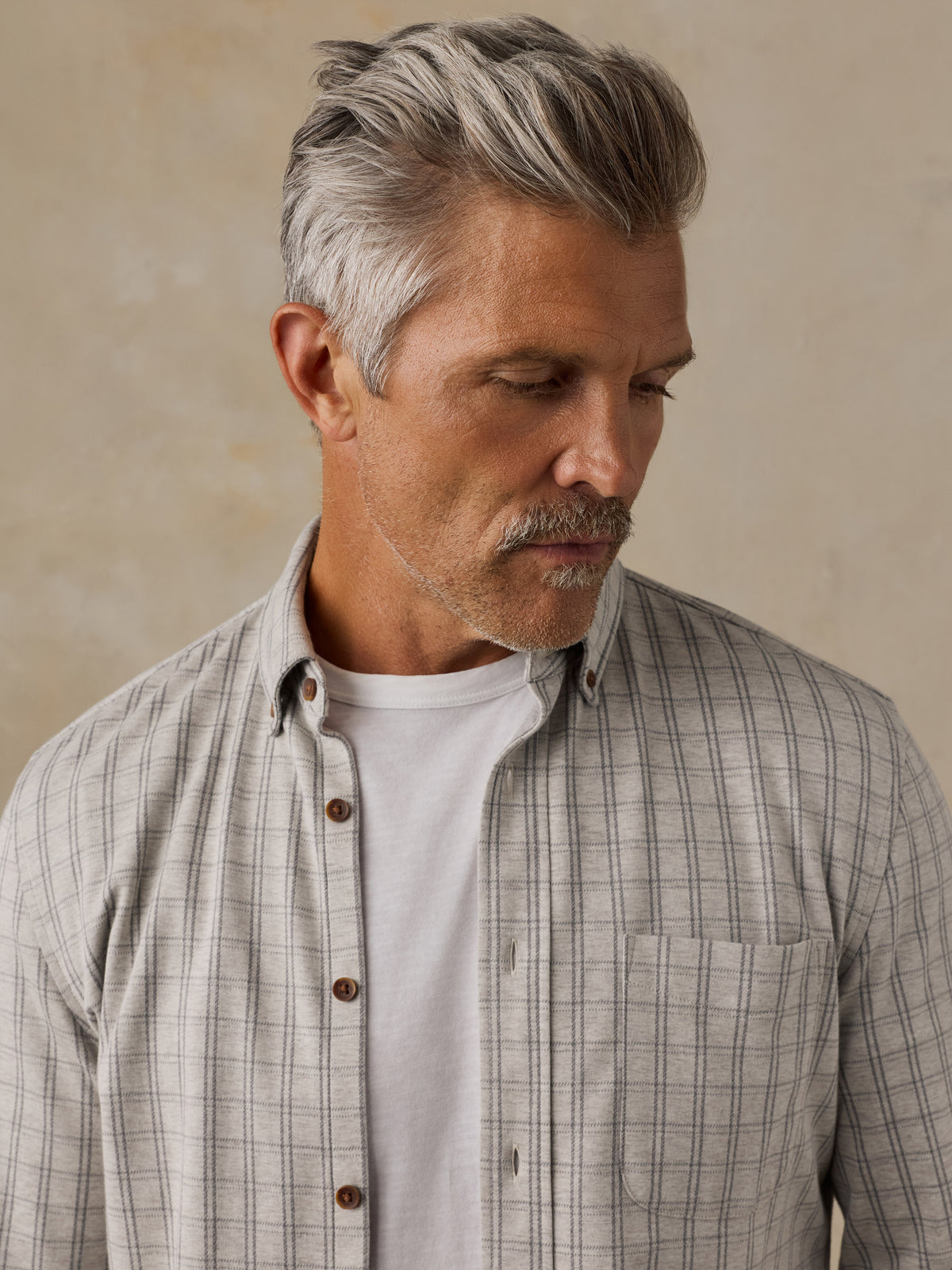 A close-up of a man wearing a light beige and gray plaid button-down shirt over a white t-shirt, set against a neutral textured beige background., image 2 of 6