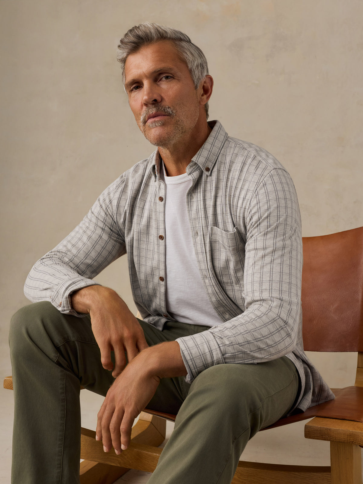 A man with gray hair and a mustache sits on a wooden chair wearing a light gray and white plaid button-down shirt over a white t-shirt and olive green pants, set against a neutral beige wall., image 5 of 6