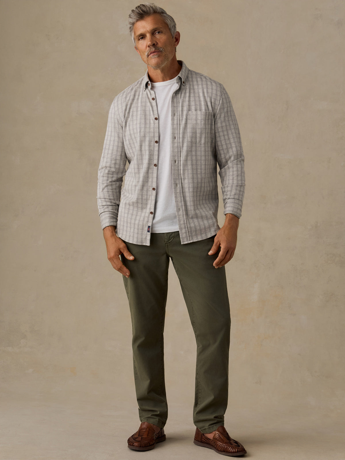 A man wearing a light gray and white checkered long-sleeve button-down shirt over a white t-shirt, paired with olive green pants and brown leather loafers, standing against a textured beige studio backdrop., image 6 of 6