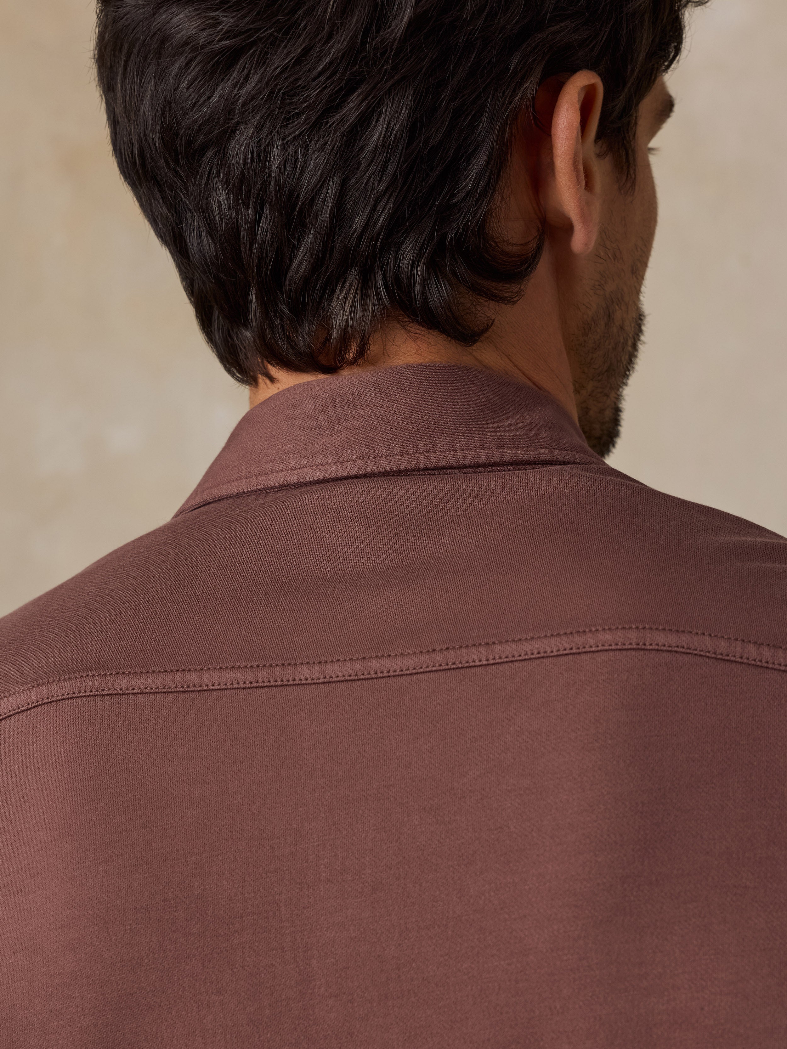 Close-up back view of a man wearing a brown collared shirt, showing the collar, upper back, and dark hair against a soft beige background.