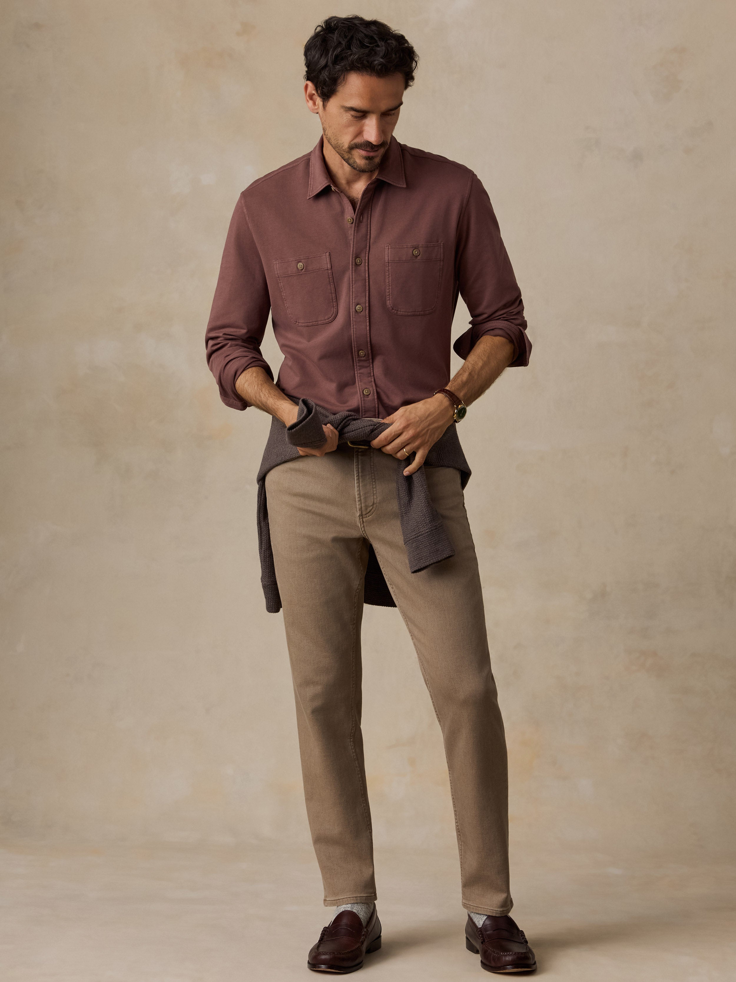 A man modeling a long-sleeve button-up shirt in a dusty rose-brown color, paired with beige trousers and dark brown loafers, standing against a textured beige studio backdrop.