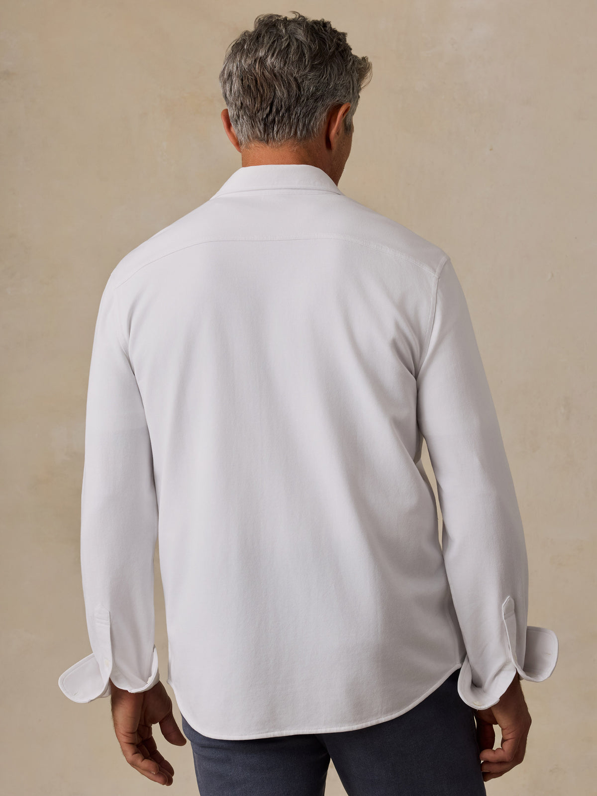 A man viewed from behind wearing a white long-sleeve button-down shirt and dark trousers, standing against a textured beige wall.