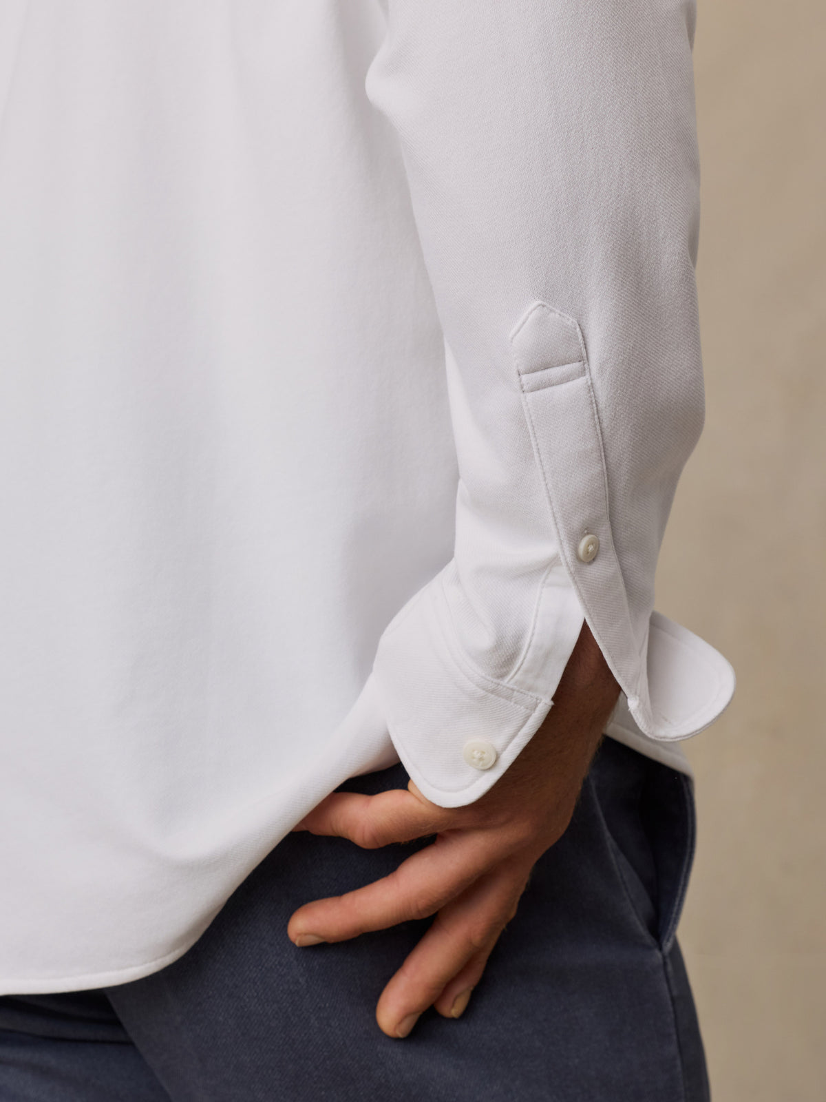 Close-up of a person wearing a white long-sleeve button-up shirt with the cuff turned up, paired with dark navy trousers, against a neutral beige background.