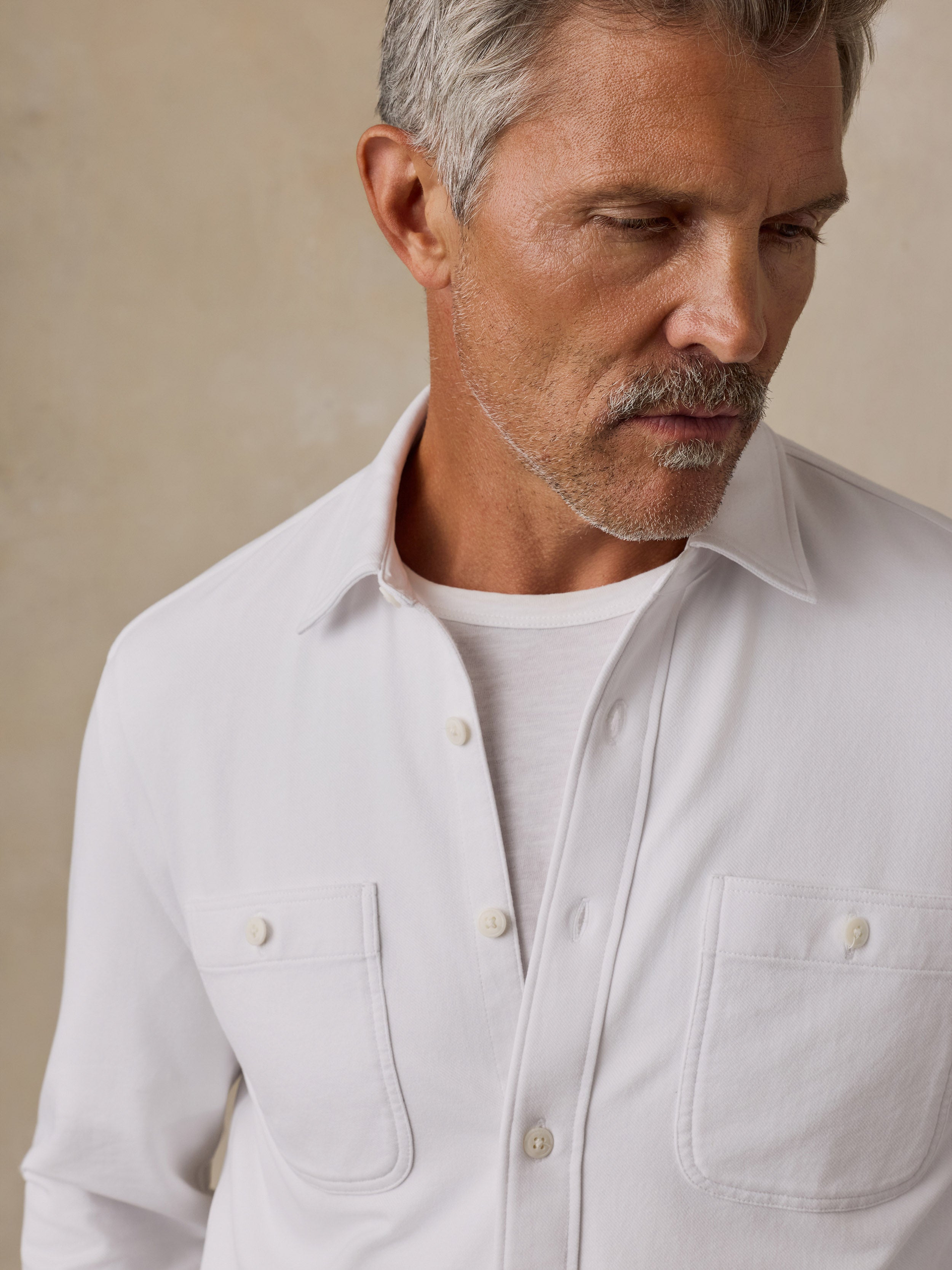 A close-up of a man wearing a white long-sleeve button-up shirt over a white t-shirt, set against a neutral beige background.