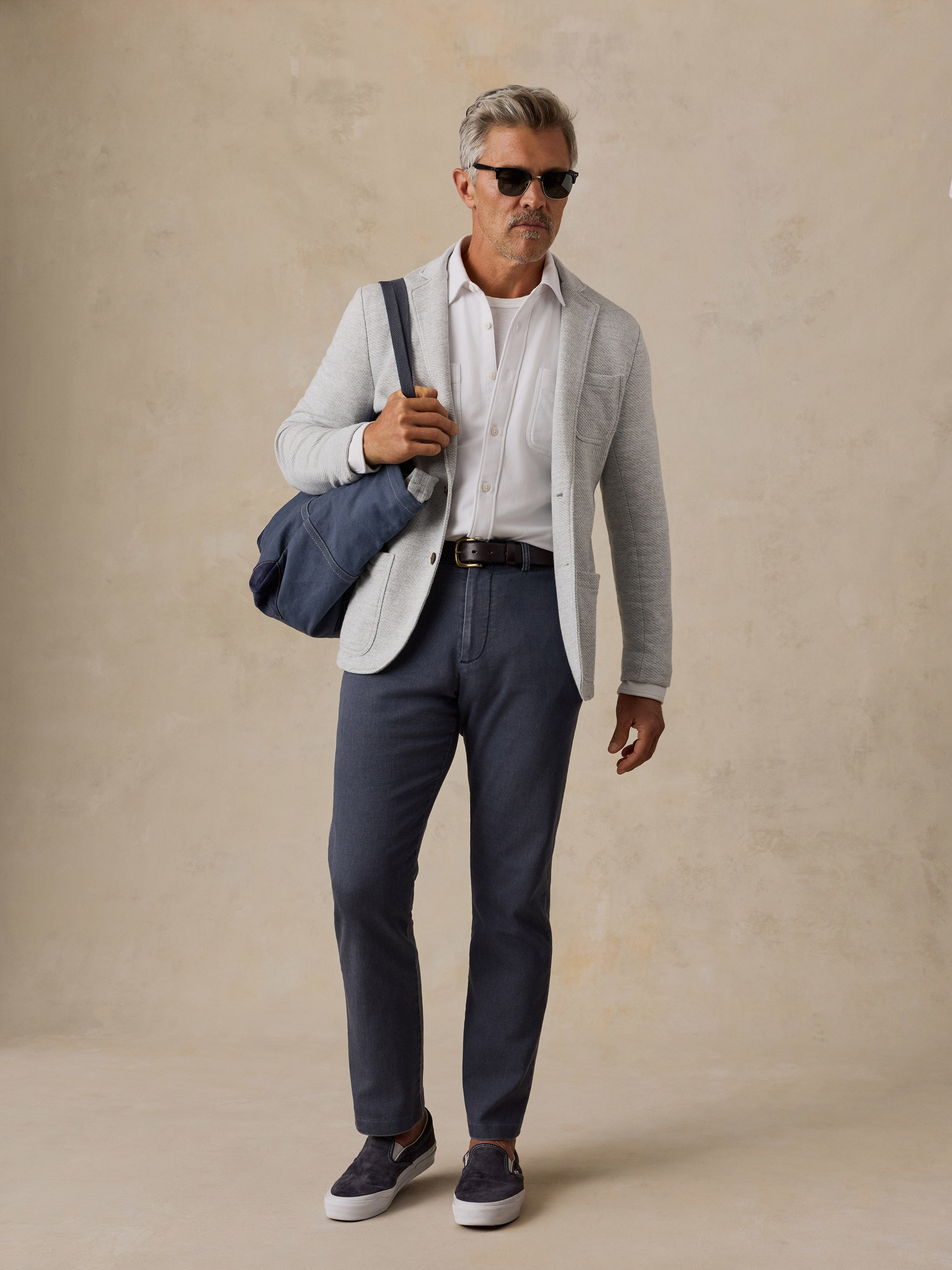 A man wearing a light gray blazer, white dress shirt, navy trousers, and navy slip-on shoes, carrying a navy duffel bag over his shoulder against a textured beige studio backdrop.