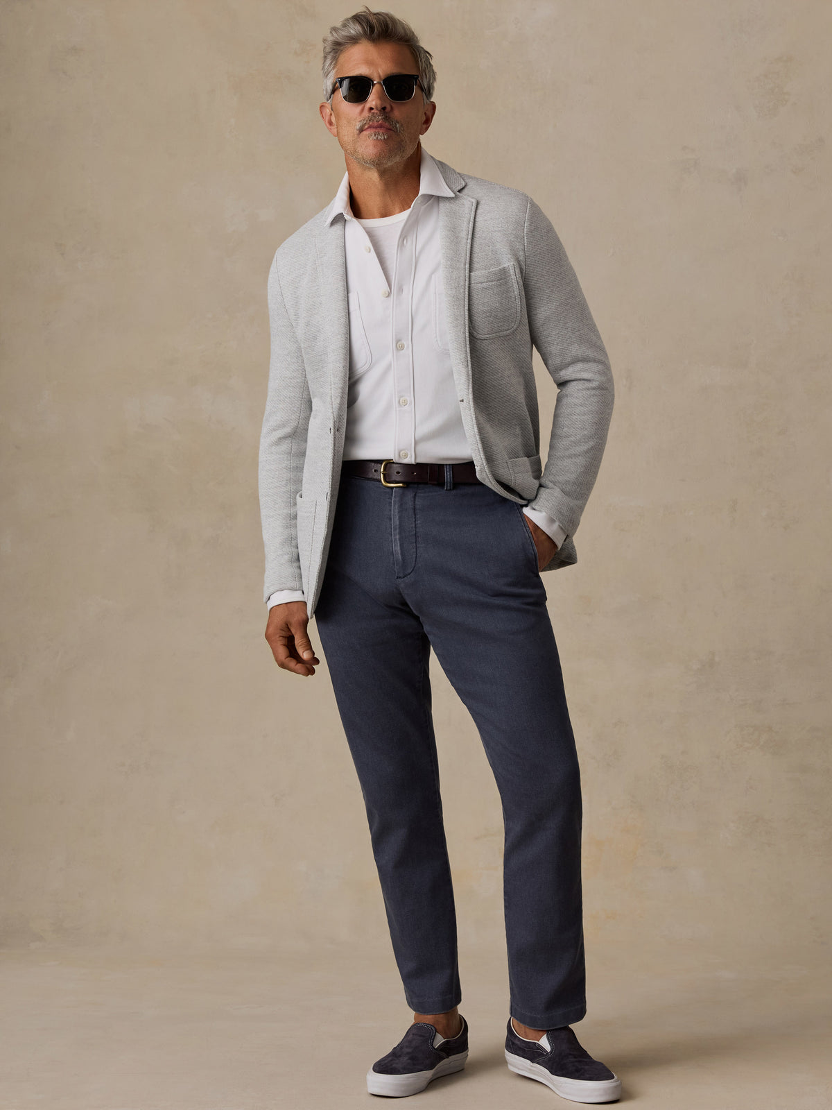 A man wearing a light gray knit cardigan over a white button-down shirt, navy blue trousers, and dark slip-on sneakers, standing against a textured beige studio backdrop.