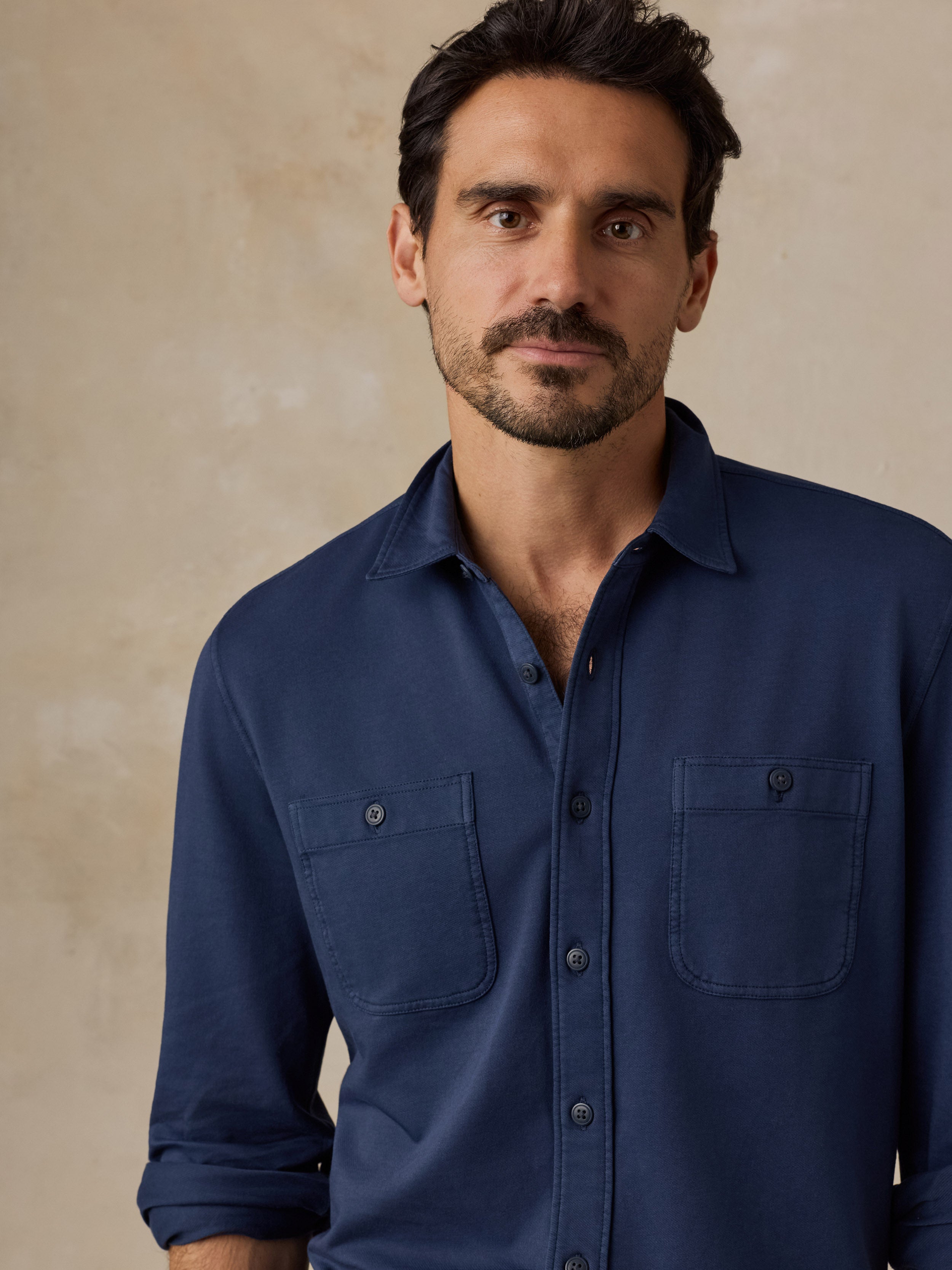 A man wearing a navy blue long-sleeve button-up shirt with two chest pockets, photographed against a textured beige background.
