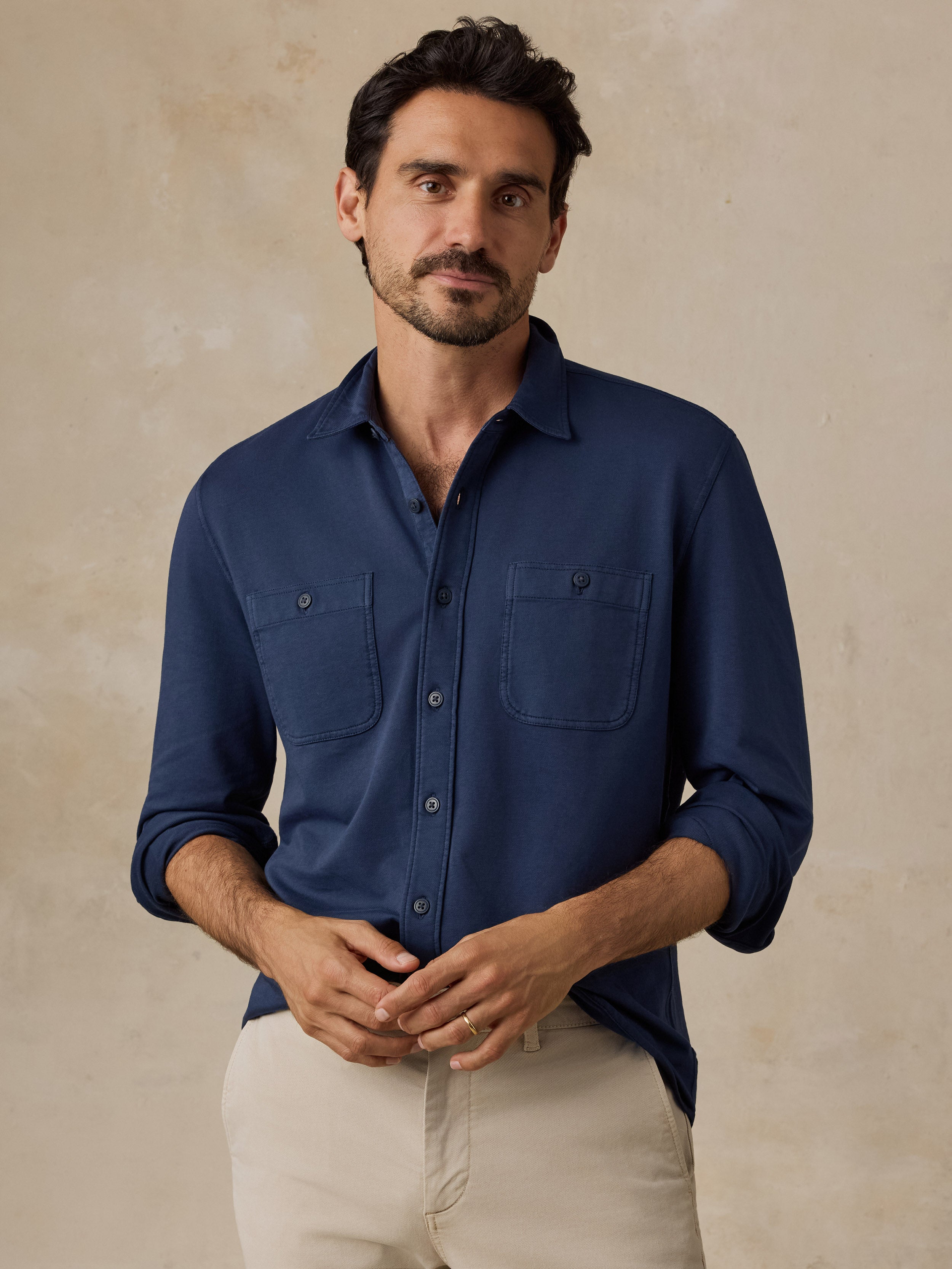 A man wearing a navy blue long-sleeve button-down shirt with two chest pockets and beige trousers, standing against a textured beige background.