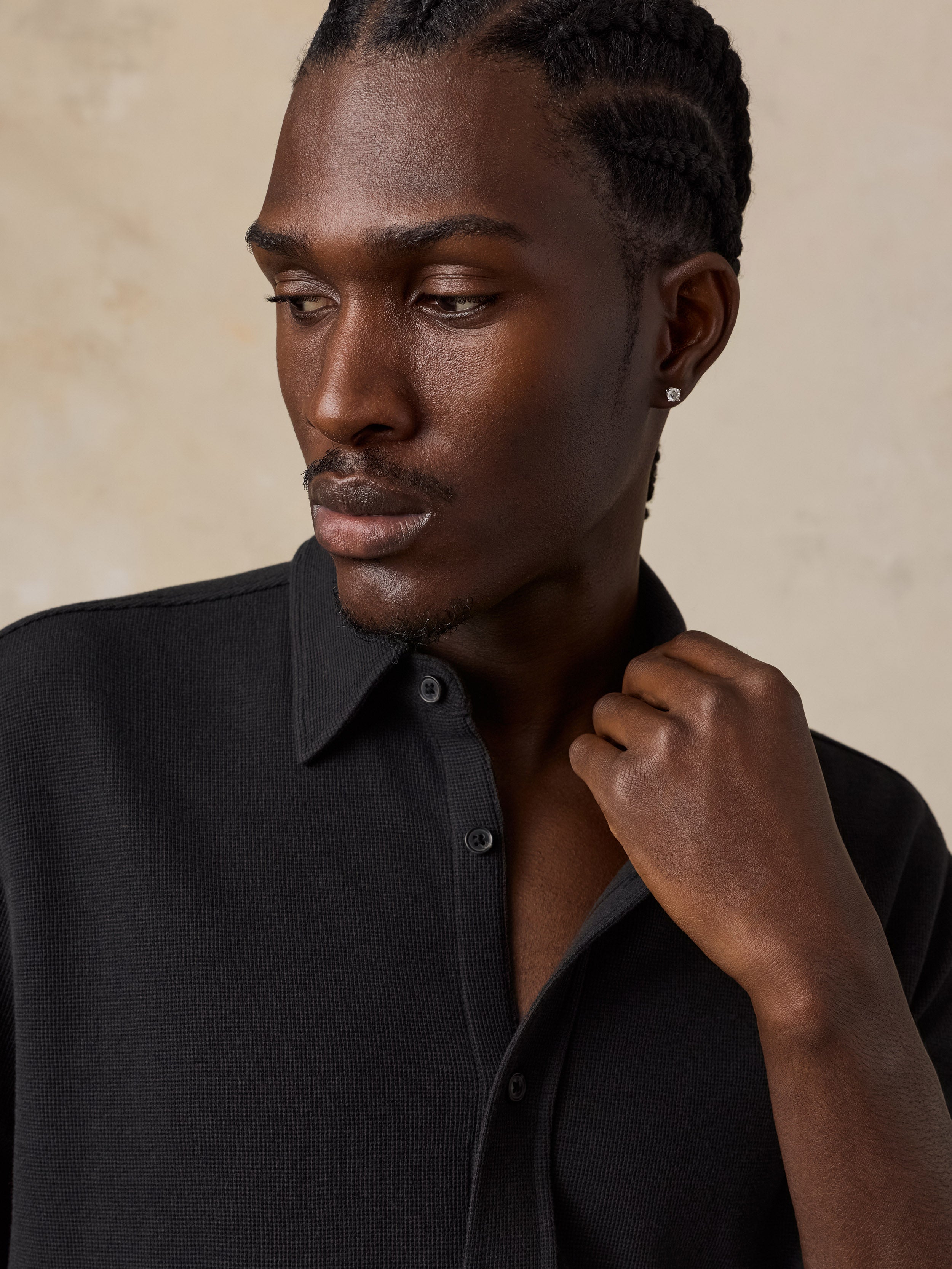 A close-up shot of a man wearing a black textured knit button-down shirt, looking down and to the side against a warm beige textured background.
