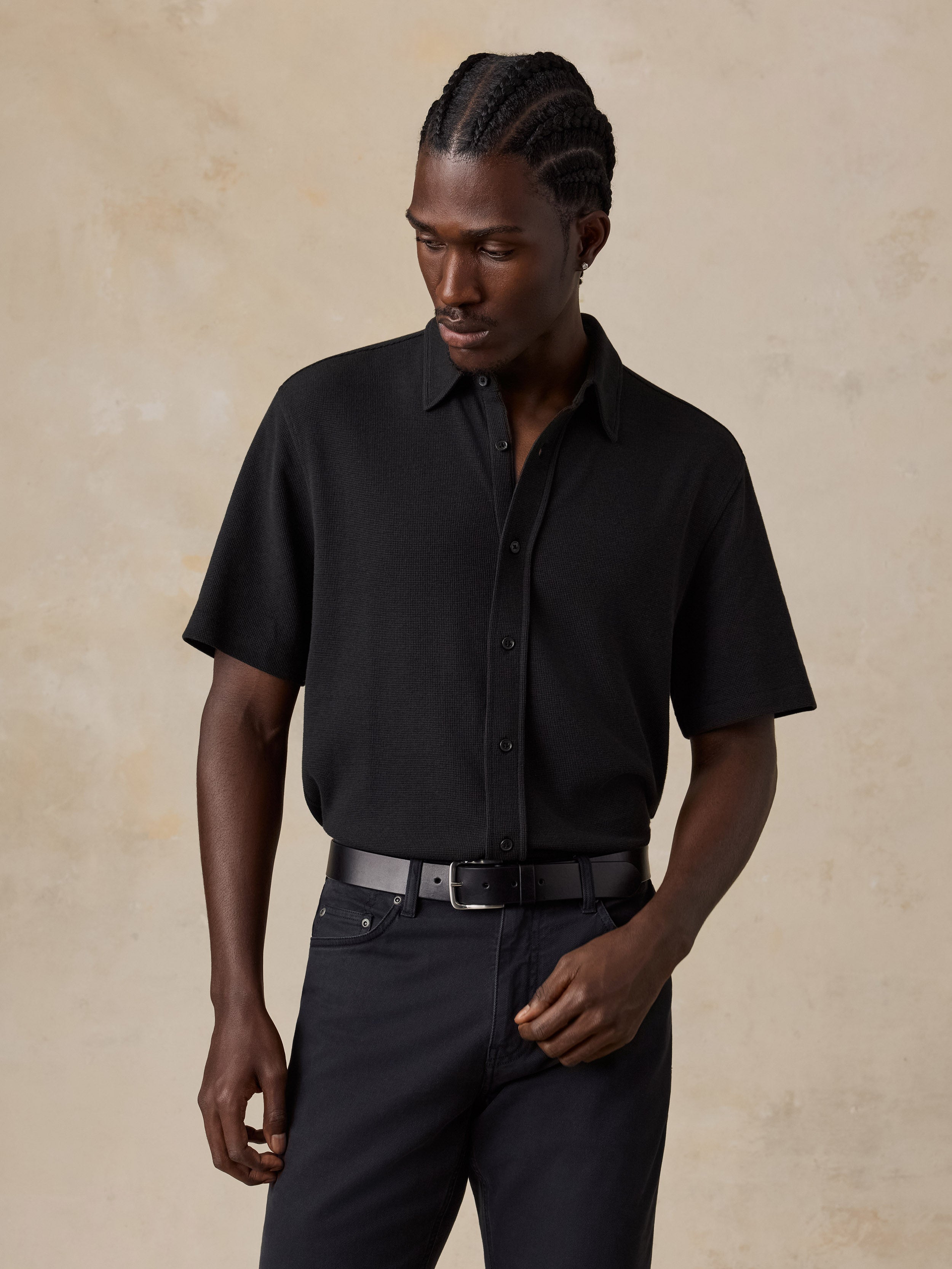 A man wearing a black short-sleeve button-up shirt and dark trousers, standing against a textured beige studio background.