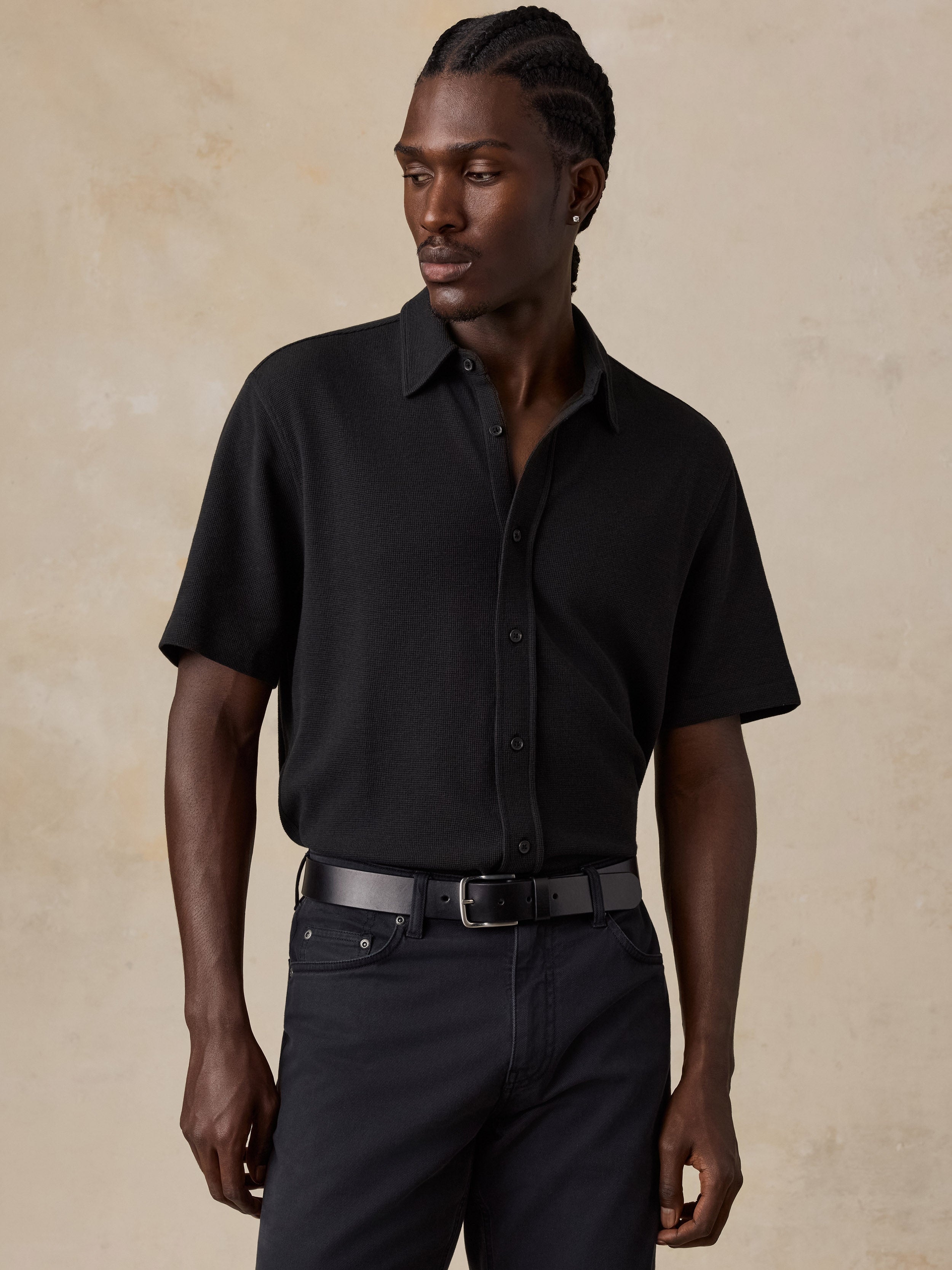 A man wearing a black short-sleeve button-up shirt tucked into black trousers with a black belt, standing against a textured beige studio background.