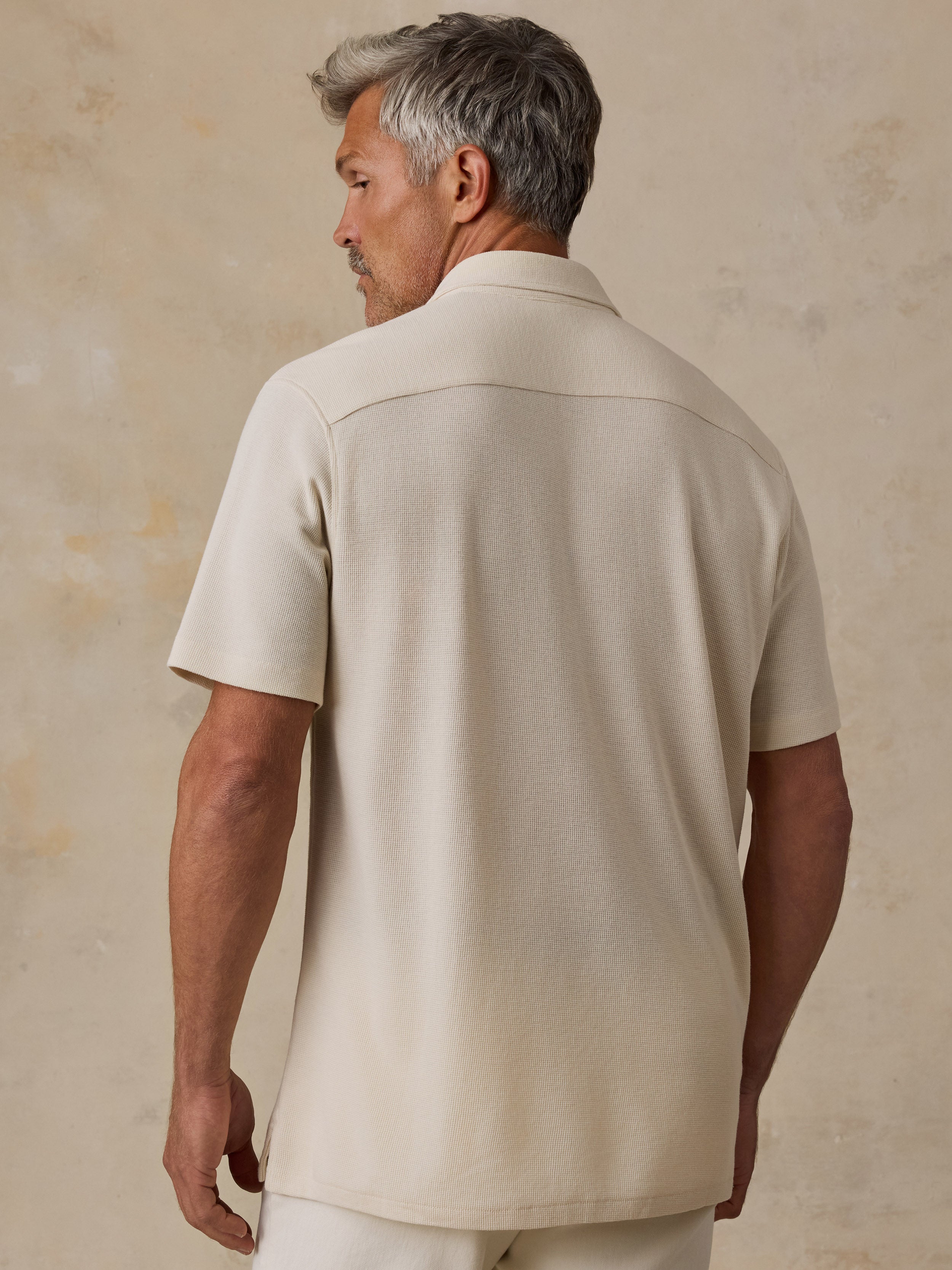 A man viewed from the back wearing a short-sleeve camp collar shirt in a warm cream or off-white color, set against a textured beige studio backdrop.