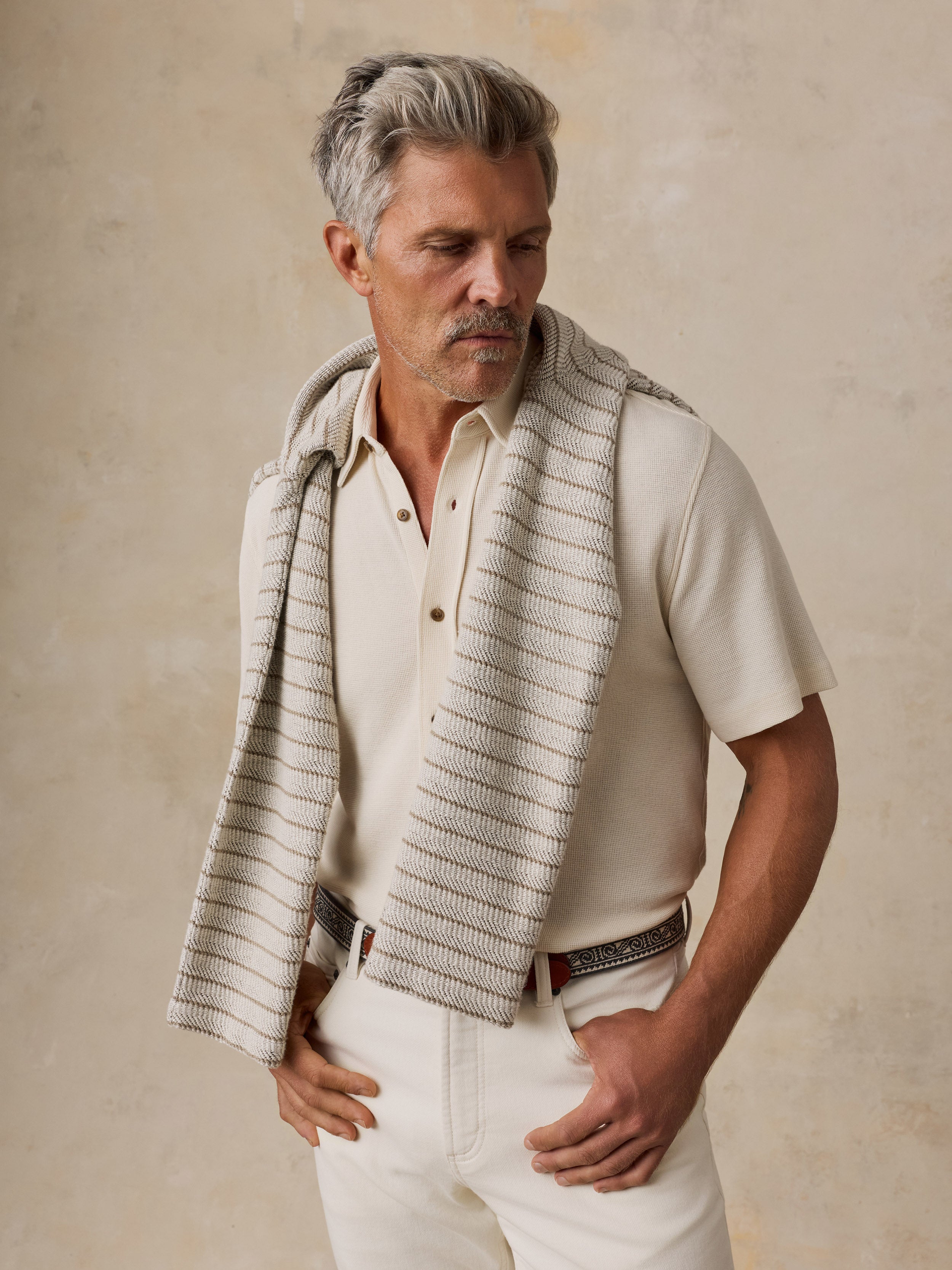 A man with gray hair and a mustache wearing a cream short-sleeve polo shirt, white trousers, and a textured cream and beige striped knit wrap draped over his shoulders, posed against a textured beige wall.