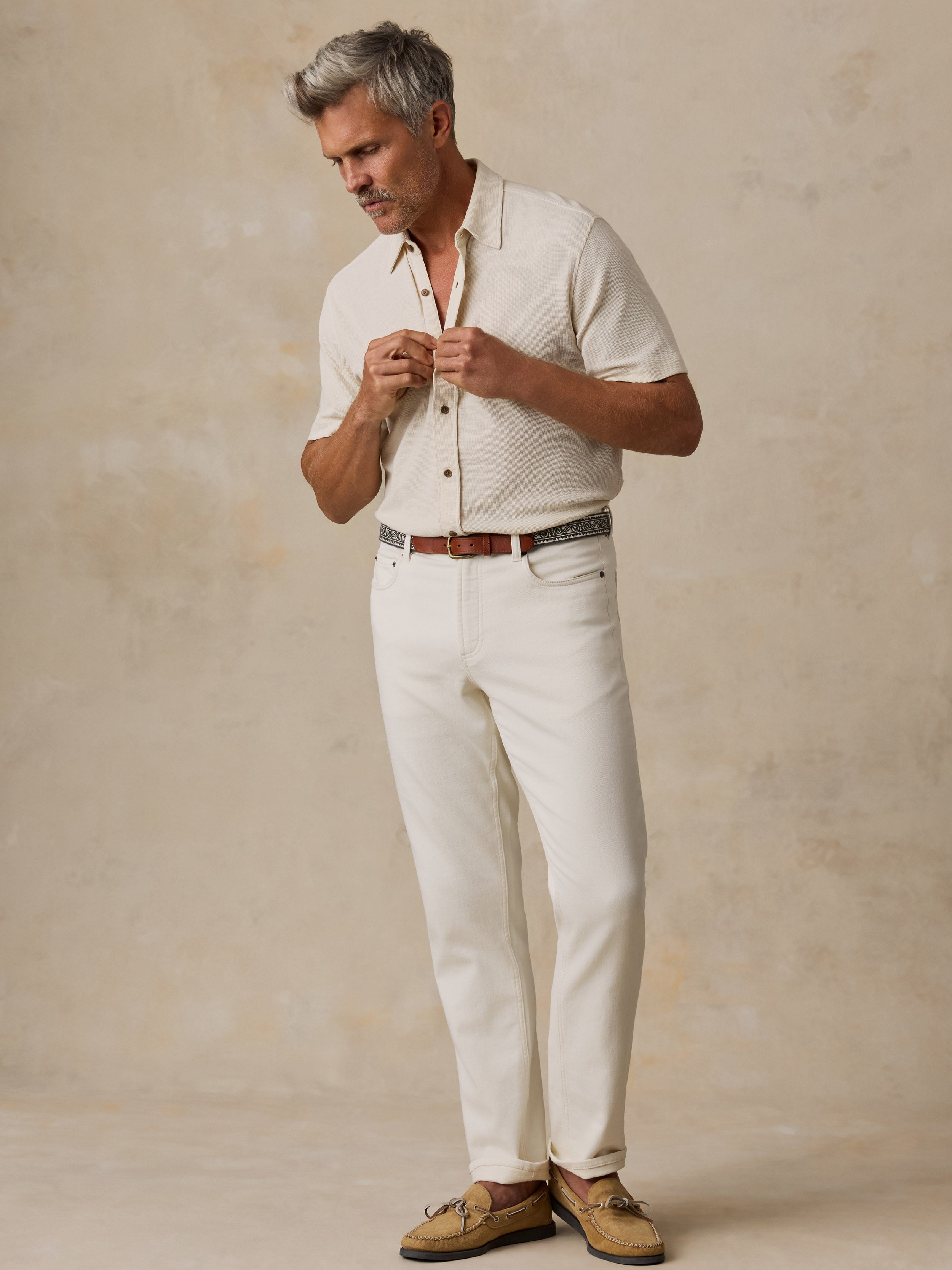A man wearing a short-sleeve cream button-up shirt, cream trousers, a brown leather belt, and tan boat shoes, standing against a textured beige studio backdrop.