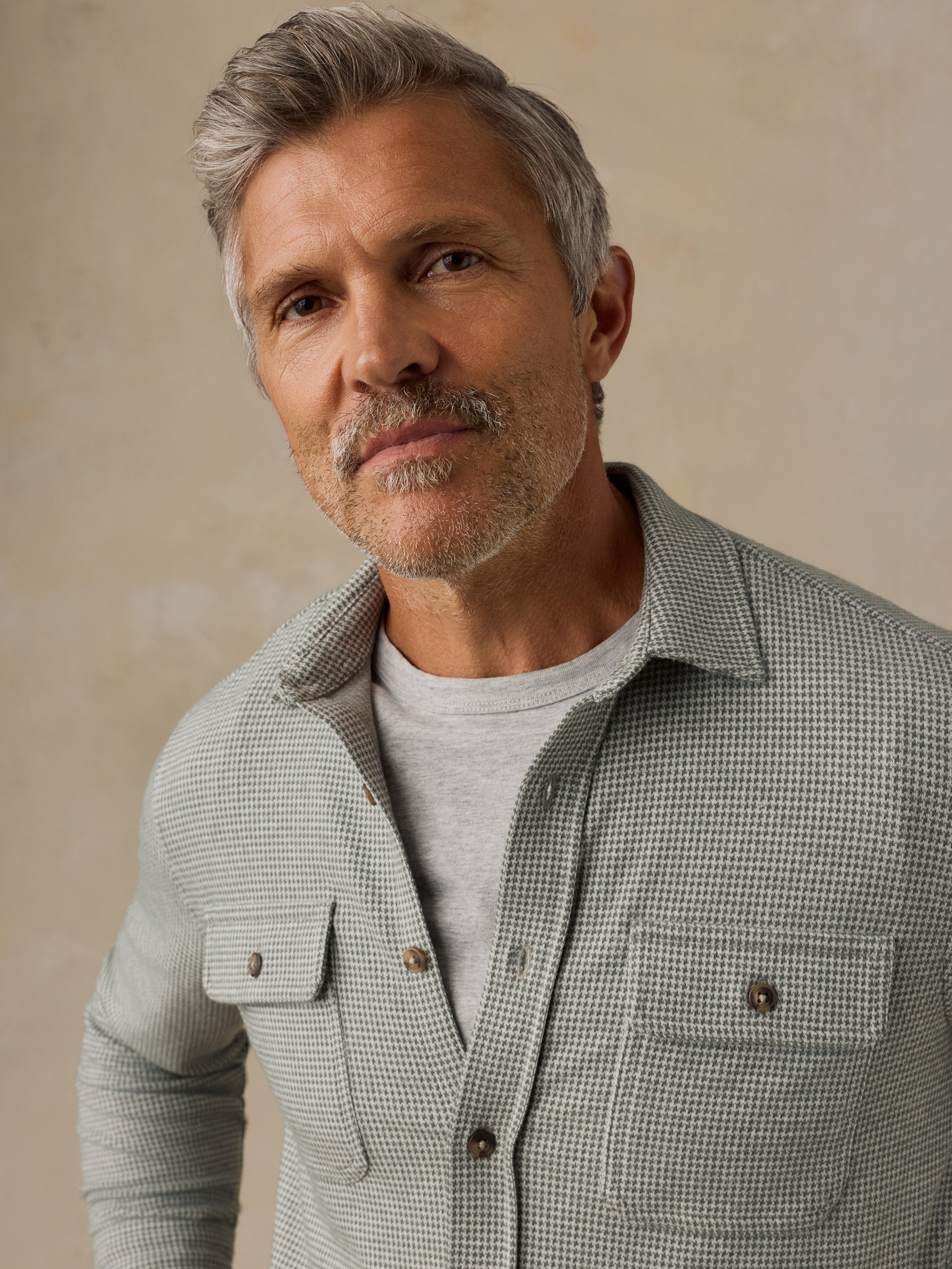A close-up portrait of a man with gray hair and a beard wearing a light sage green and white micro-check patterned button-down shirt over a light gray t-shirt, set against a neutral beige background.