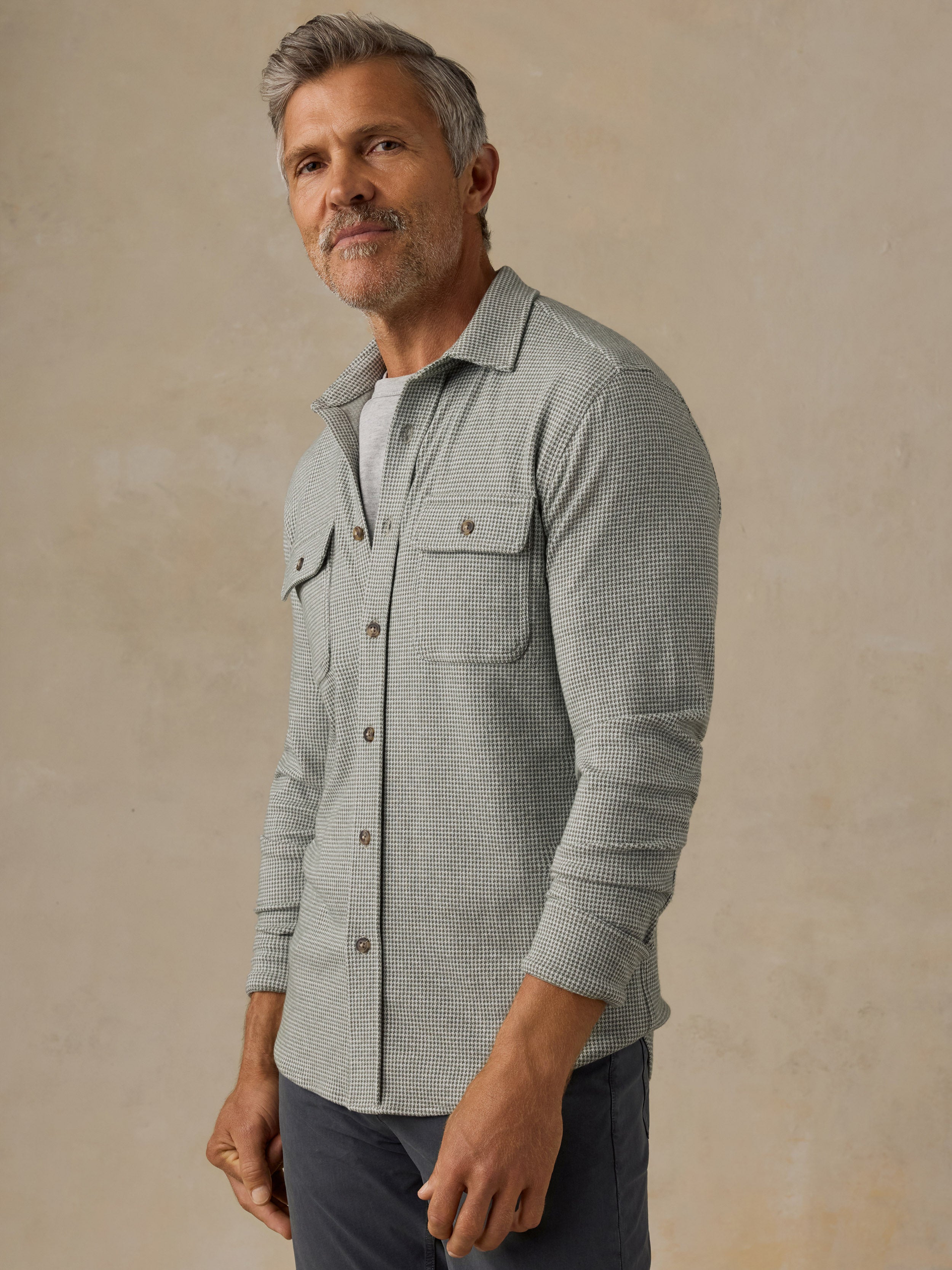A man wearing a light green and white micro-check patterned long-sleeve button-up shirt over a white undershirt, paired with dark gray pants, standing against a textured beige background.