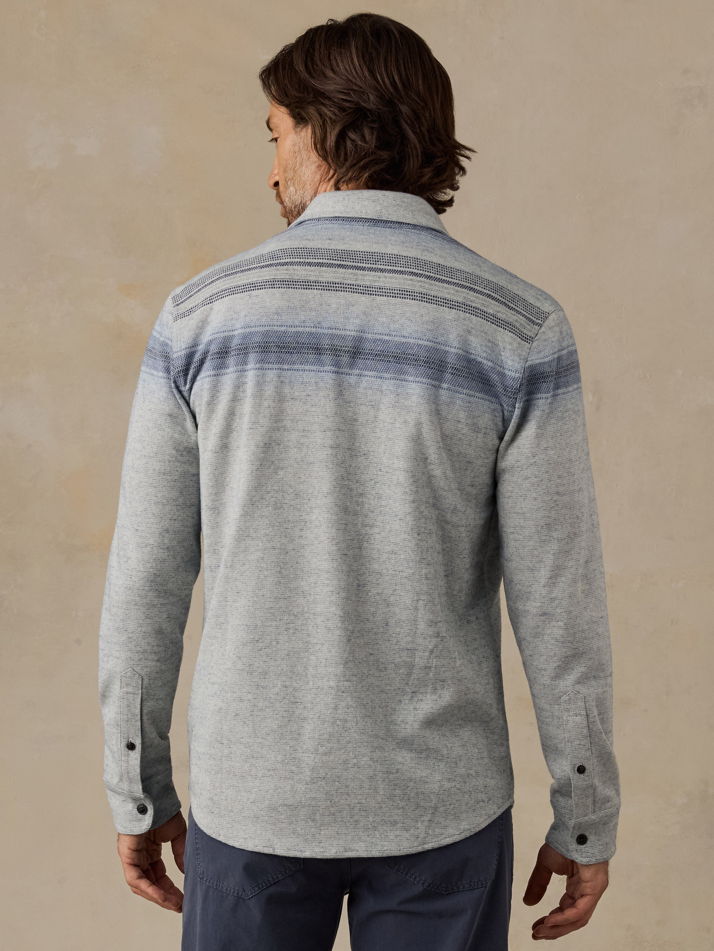 A man viewed from the back wearing a light gray long-sleeve button-up shirt with horizontal blue and gray stripes across the shoulders and upper back, paired with dark pants, against a textured beige background.