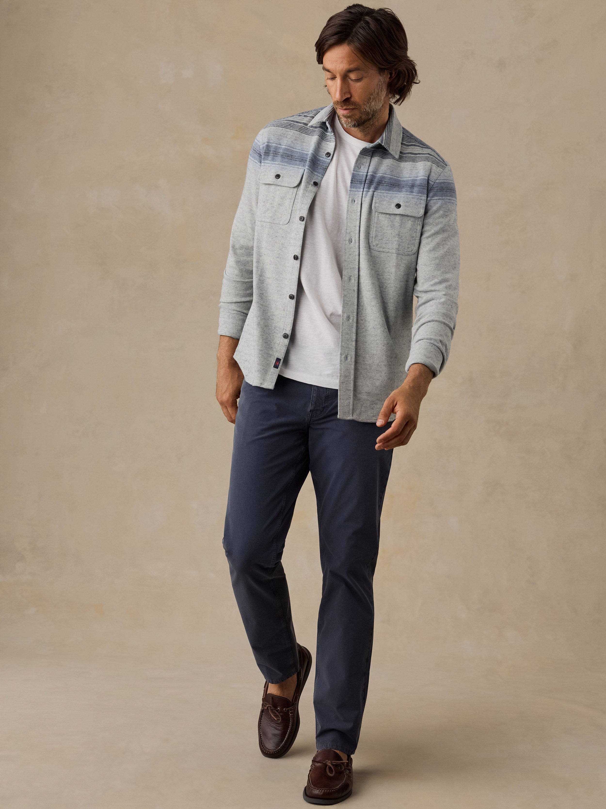 A man wearing a light gray and blue striped button-down shirt over a white t-shirt, navy blue pants, and brown leather loafers, standing against a textured beige studio backdrop.