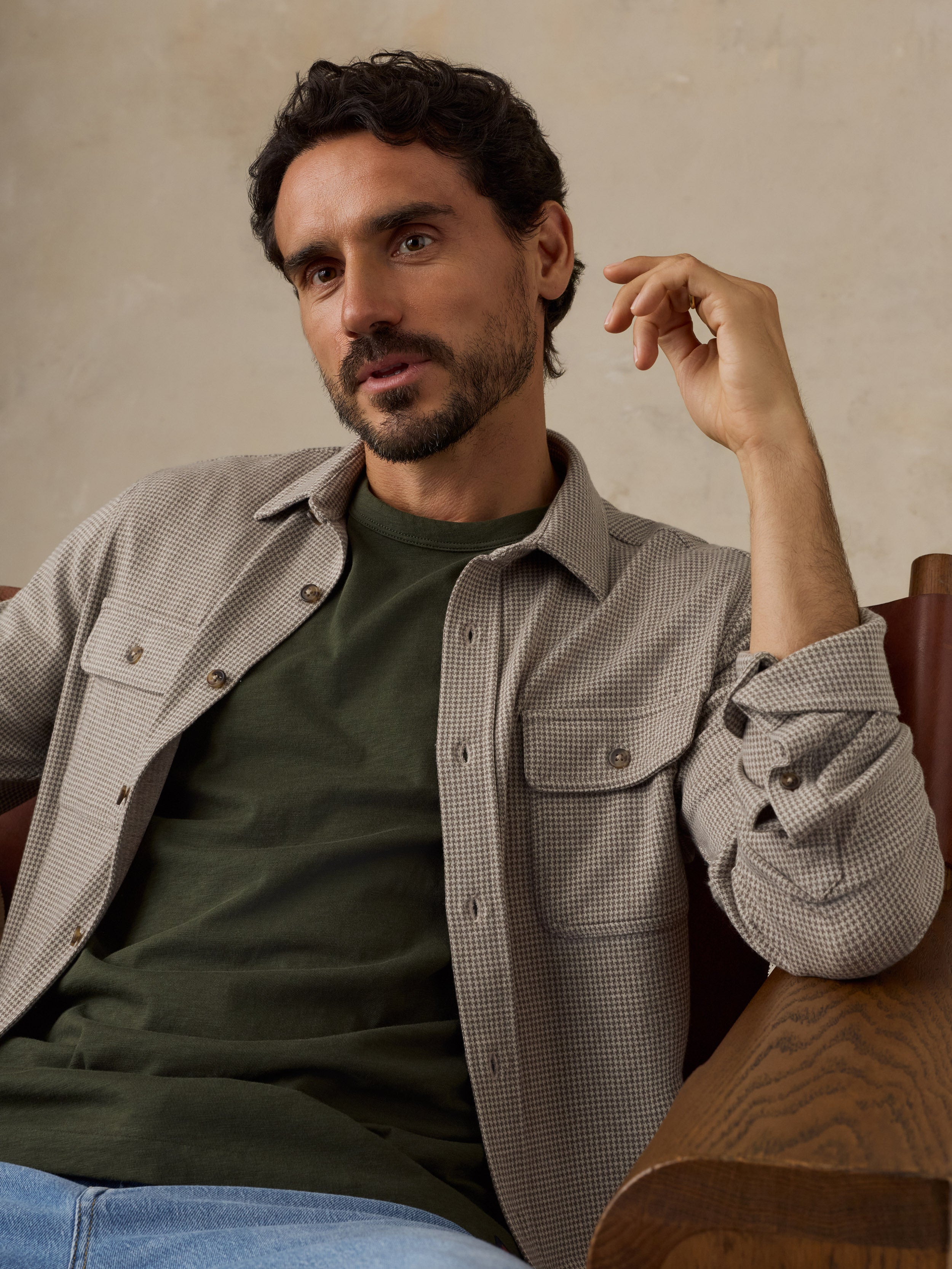 Man sitting on a wooden chair wearing a beige overshirt with a green t-shirt and jeans against a beige studio background