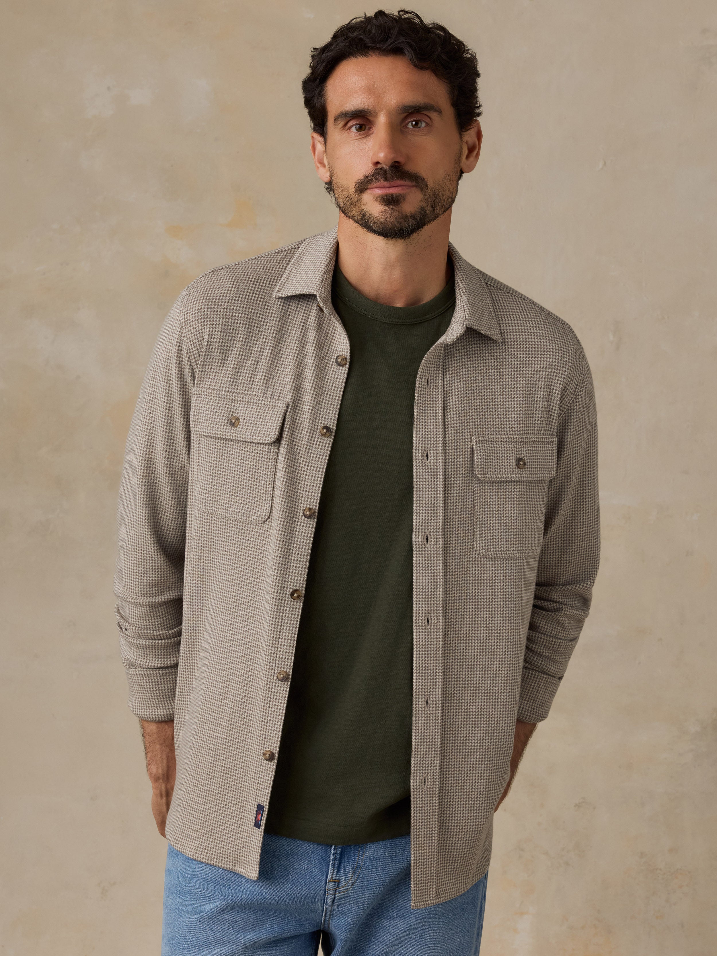 Man wearing a beige overshirt with a green t-shirt and jeans against a beige studio background