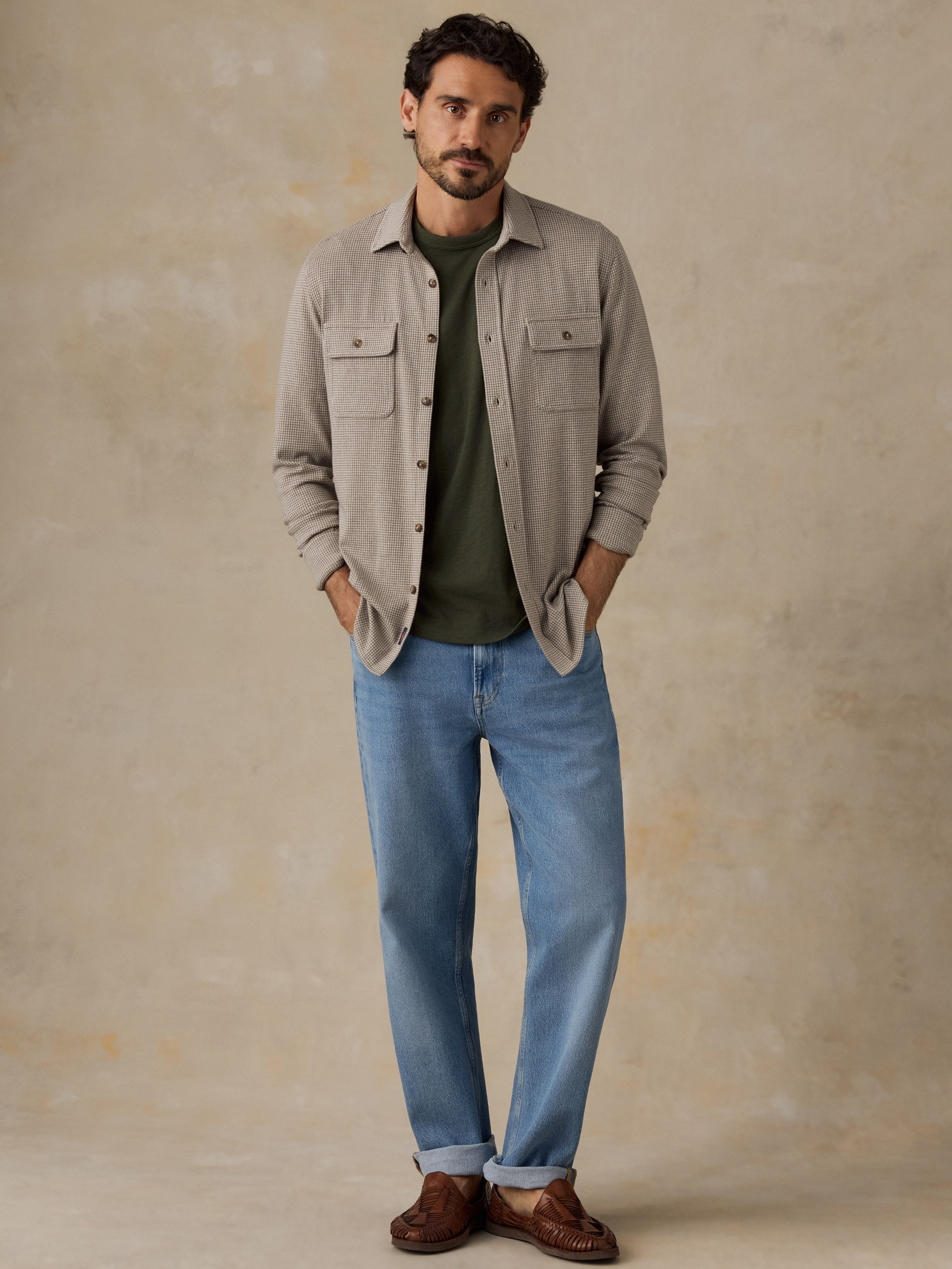 Man wearing a beige overshirt with a green t-shirt and jeans against a beige studio background