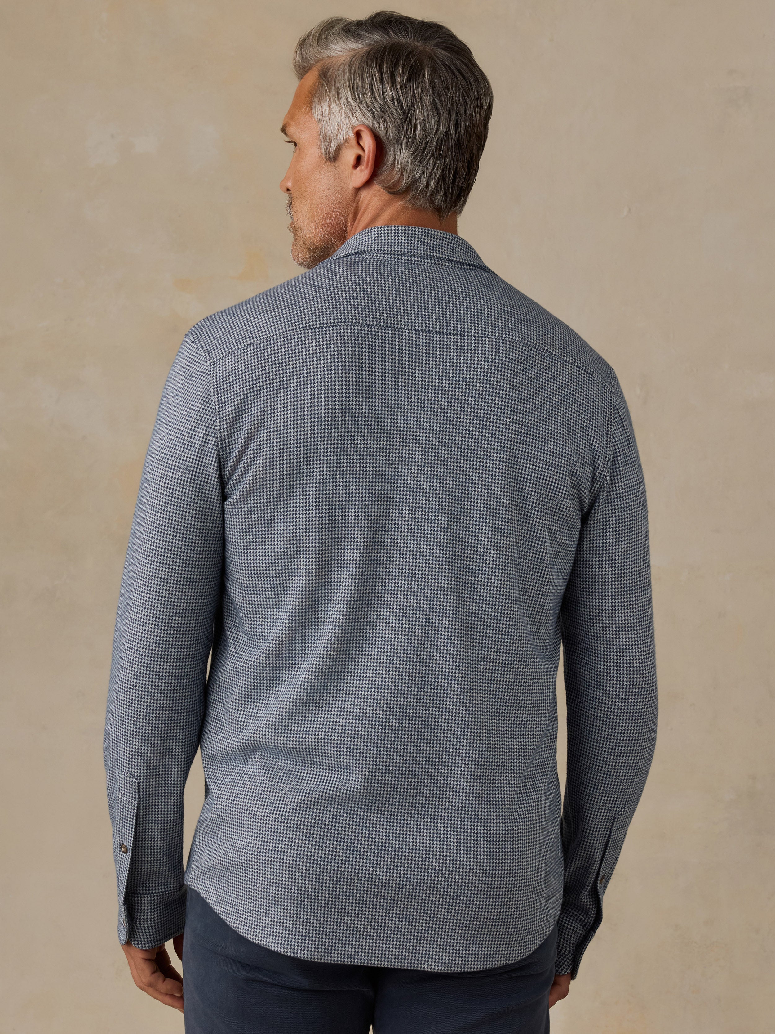 A man viewed from the back wearing a long-sleeve button-down shirt with a fine micro-check pattern in gray and white, paired with dark trousers, set against a textured beige background.