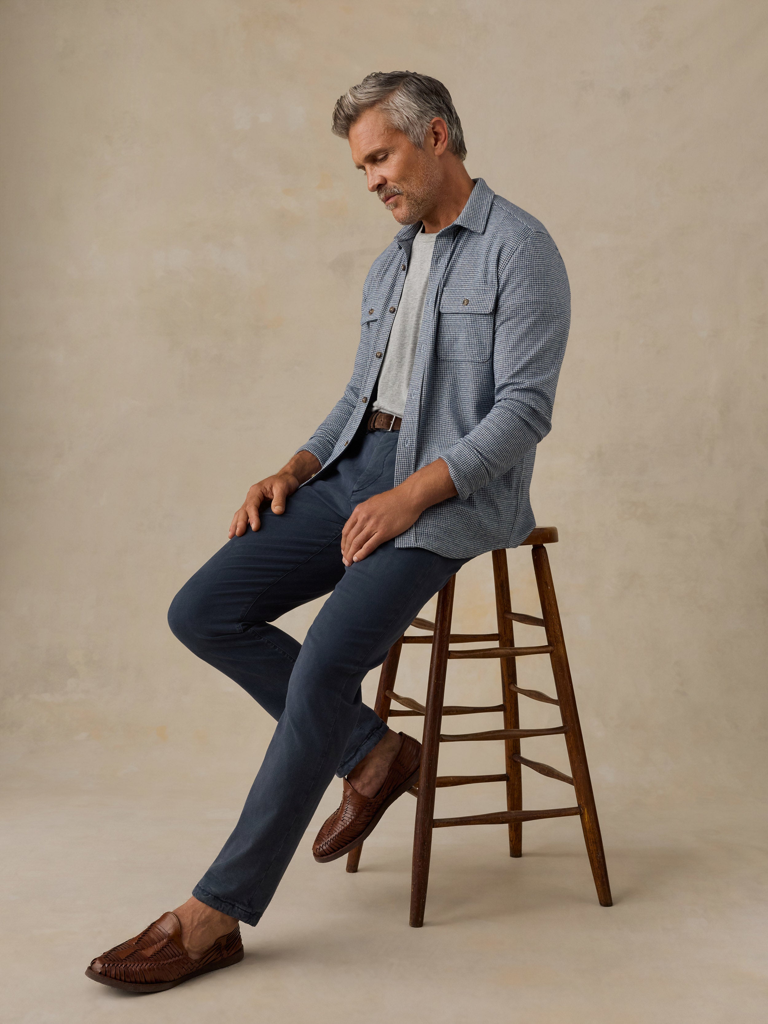 A man with gray hair sits on a wooden stool wearing a light blue denim shirt over a white t-shirt, navy blue pants, and brown leather loafers, against a textured beige studio backdrop.