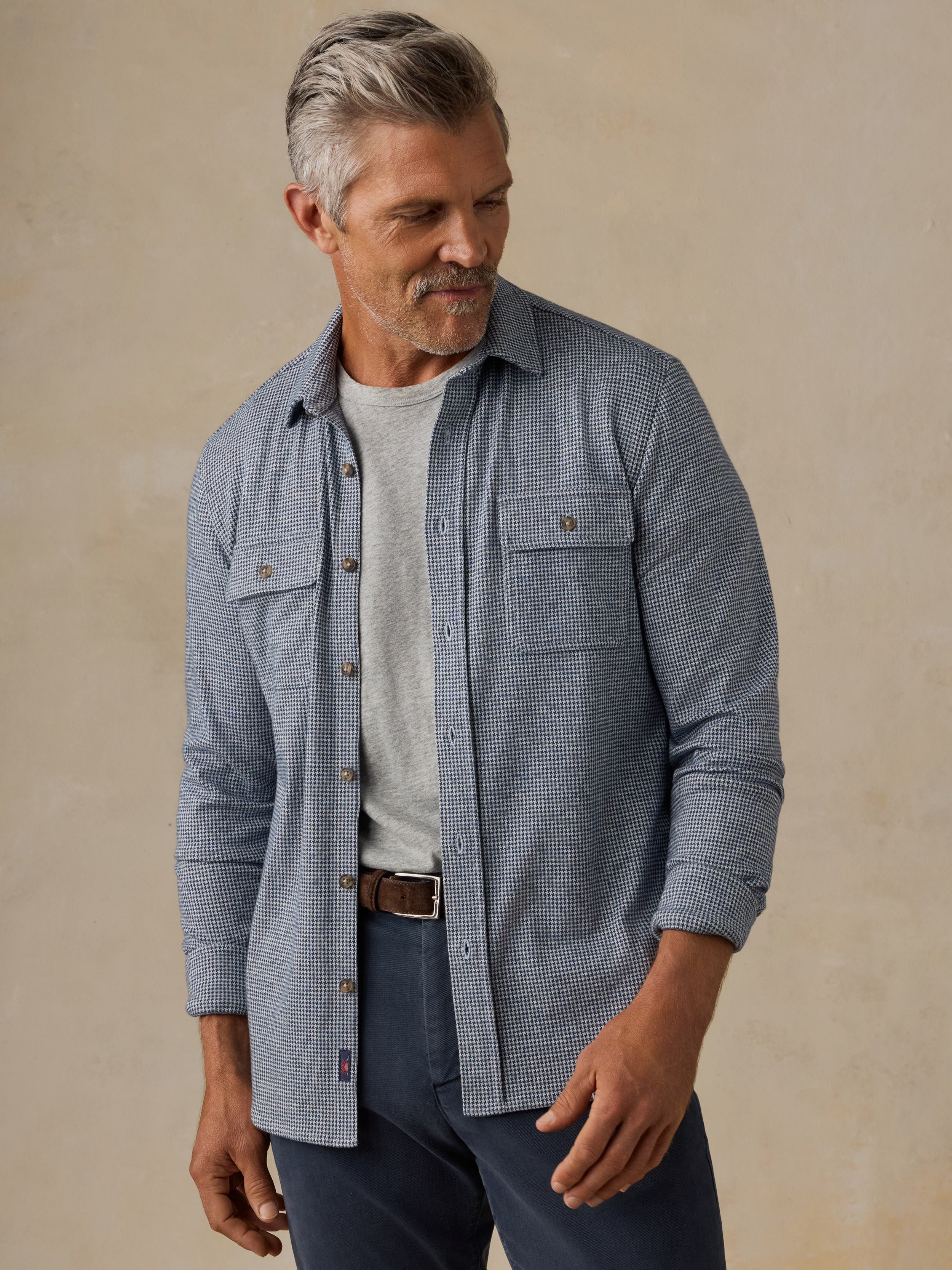 A man wearing a light blue and white micro-check patterned button-down shirt over a light gray t-shirt, paired with navy pants and a brown belt, standing against a neutral beige background.