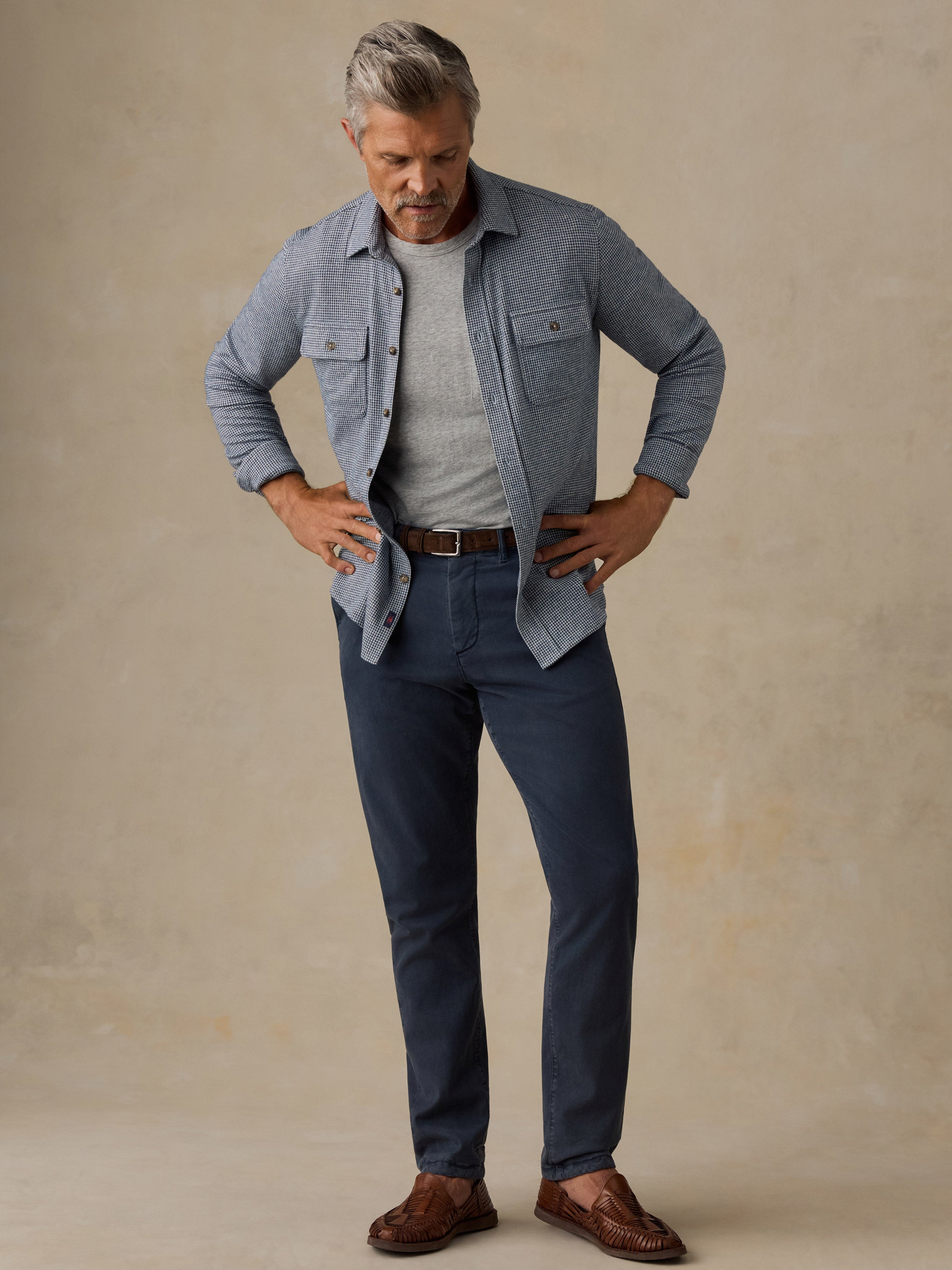 A man modeling a light blue button-up shirt over a gray t-shirt, paired with navy blue jeans, a brown belt, and brown leather loafers, standing against a textured beige studio backdrop.