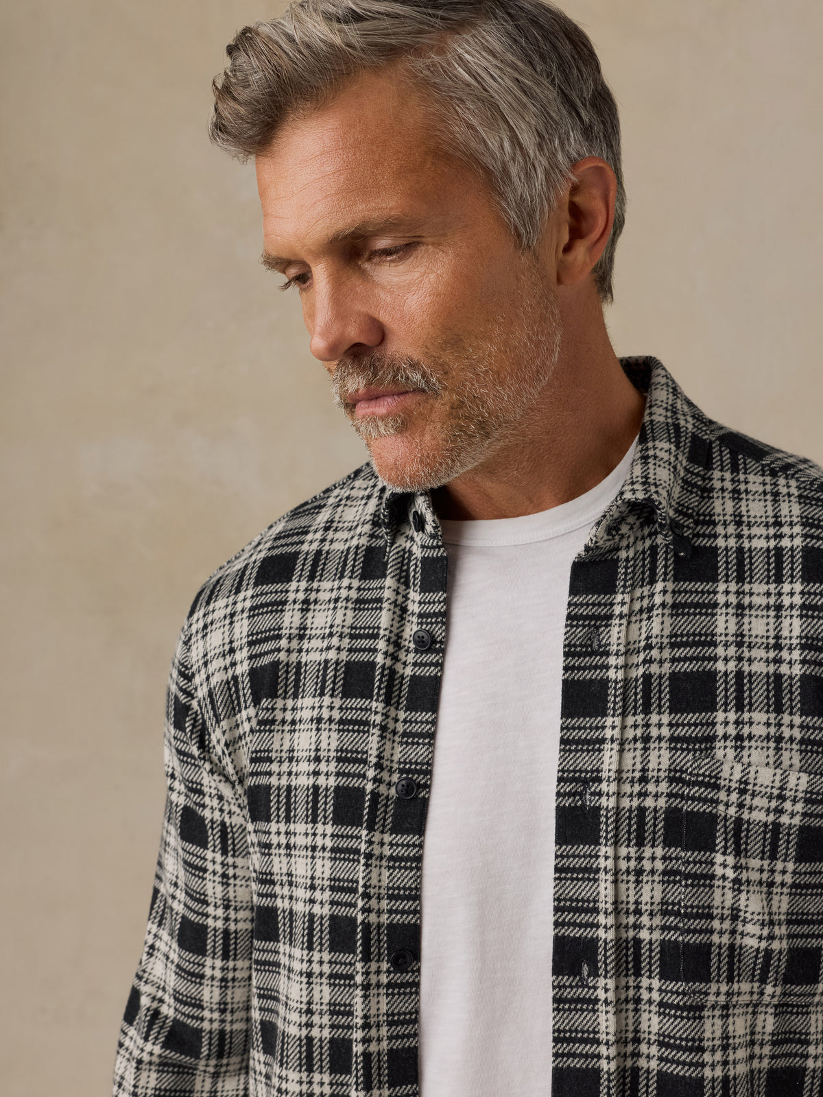 A close-up of a man wearing a black and white plaid button-down shirt over a white t-shirt, set against a neutral beige background., image 2 of 6