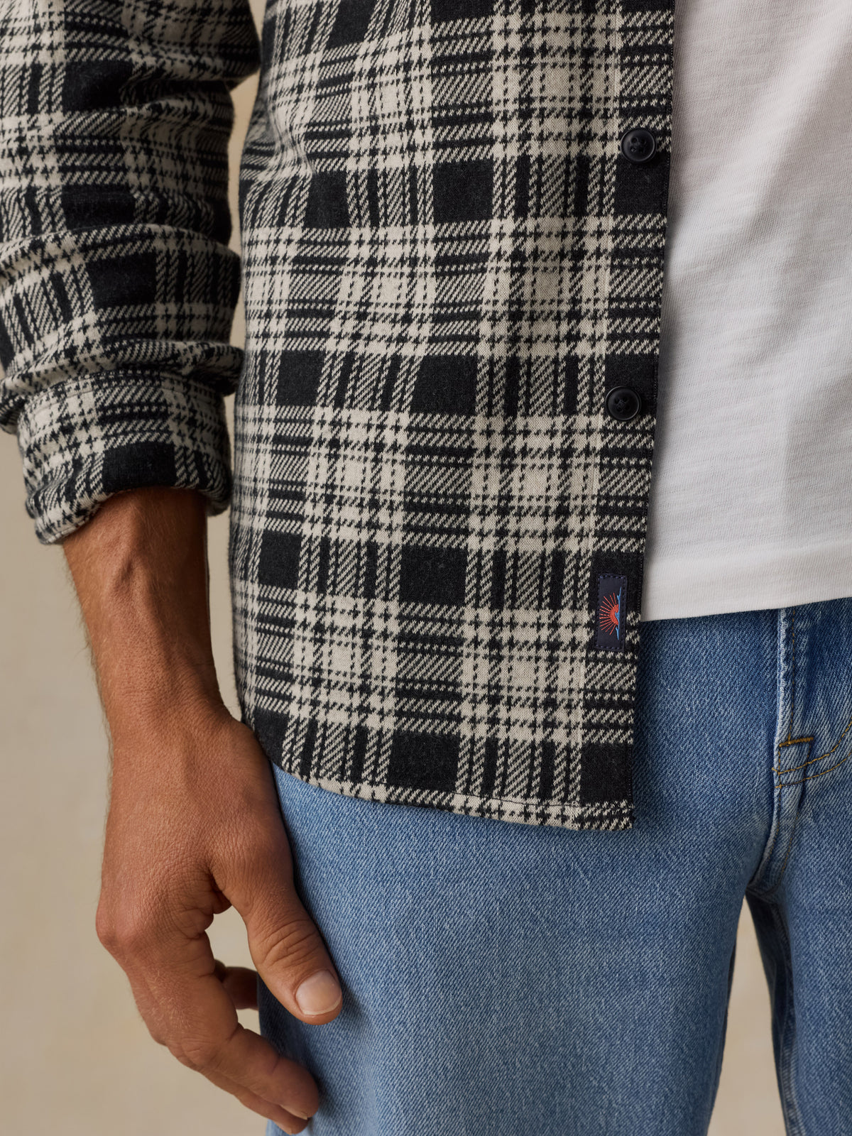 Close-up of a man wearing a black, white, and gray plaid button-up shirt over a white T-shirt and blue jeans, with a small Faherty brand tab near the hem, image 4 of 6