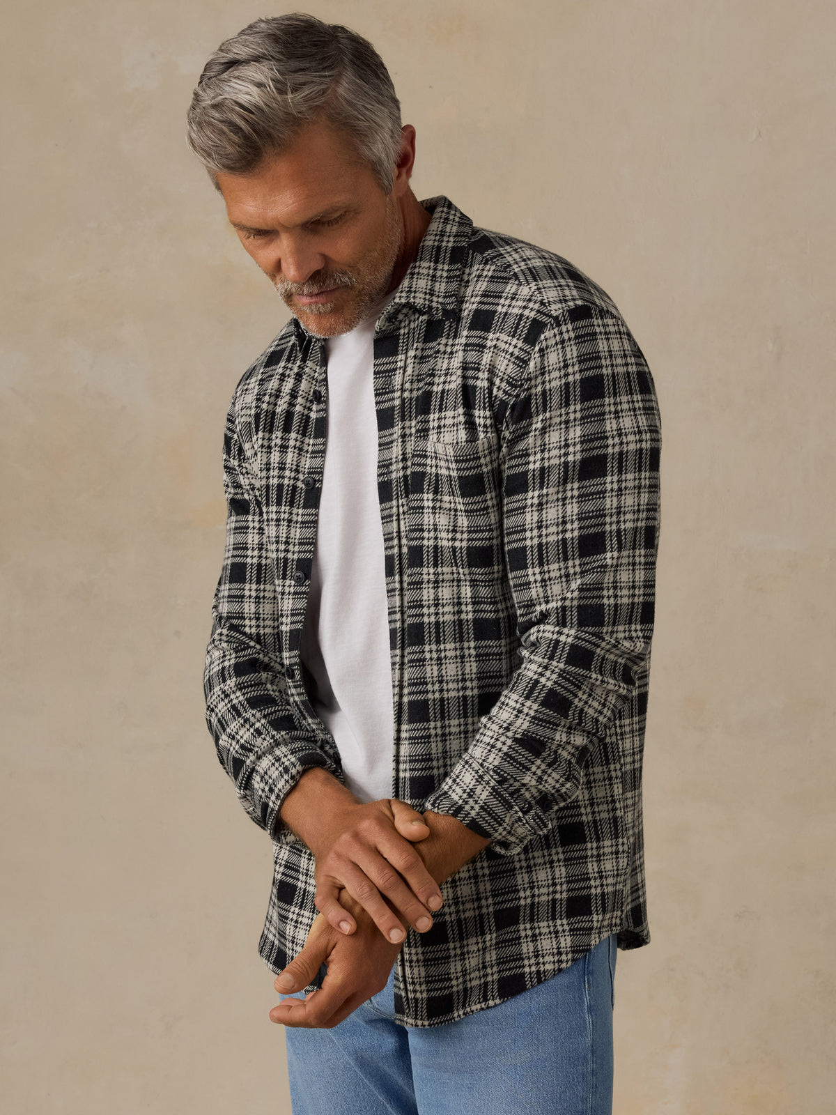 A man wearing a black and white plaid button-down shirt over a white t-shirt and blue jeans, standing against a textured beige wall., image 1 of 6