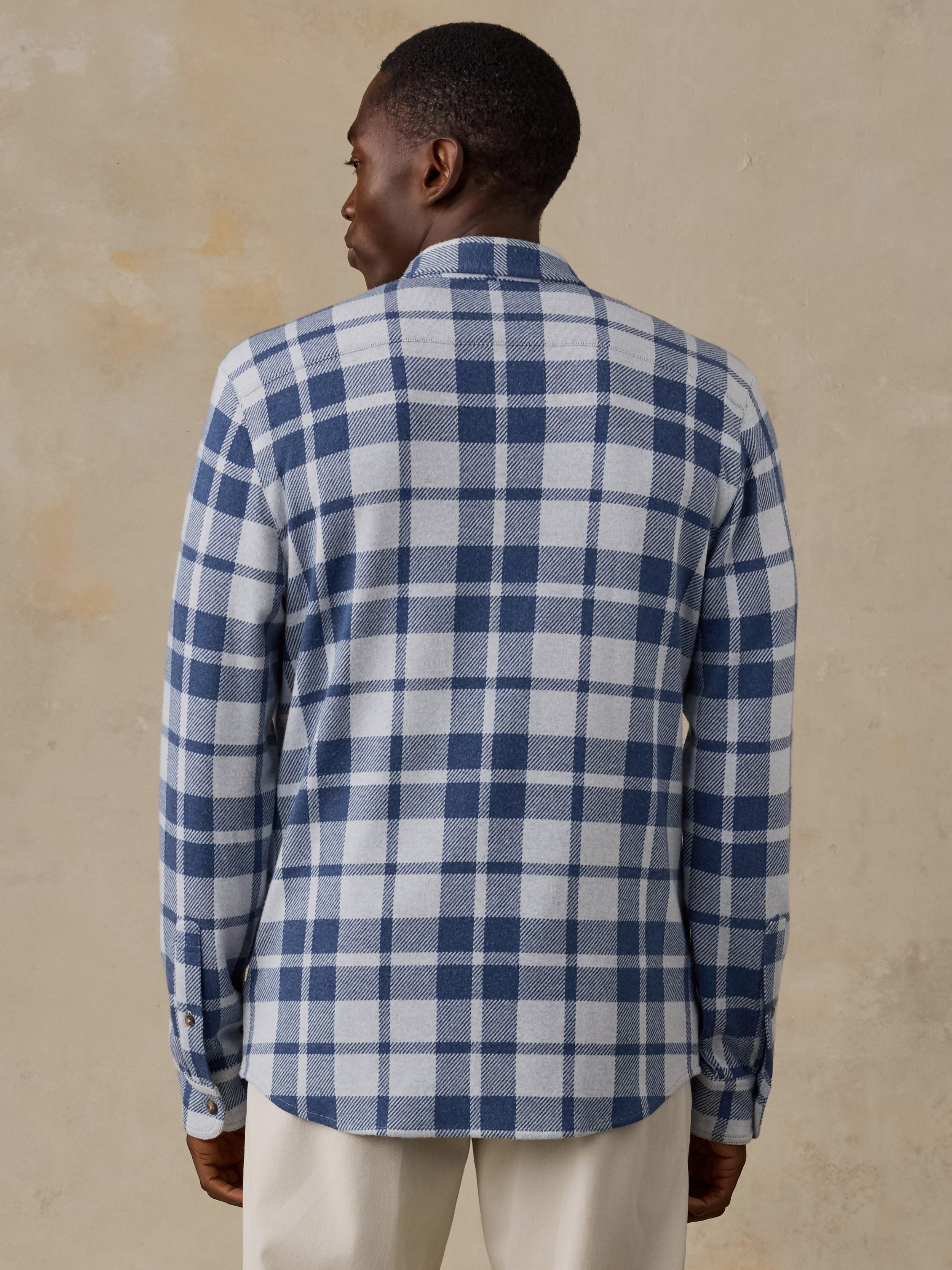 A man viewed from the back wearing a blue and white plaid long-sleeve shirt and cream trousers, standing against a textured beige wall.