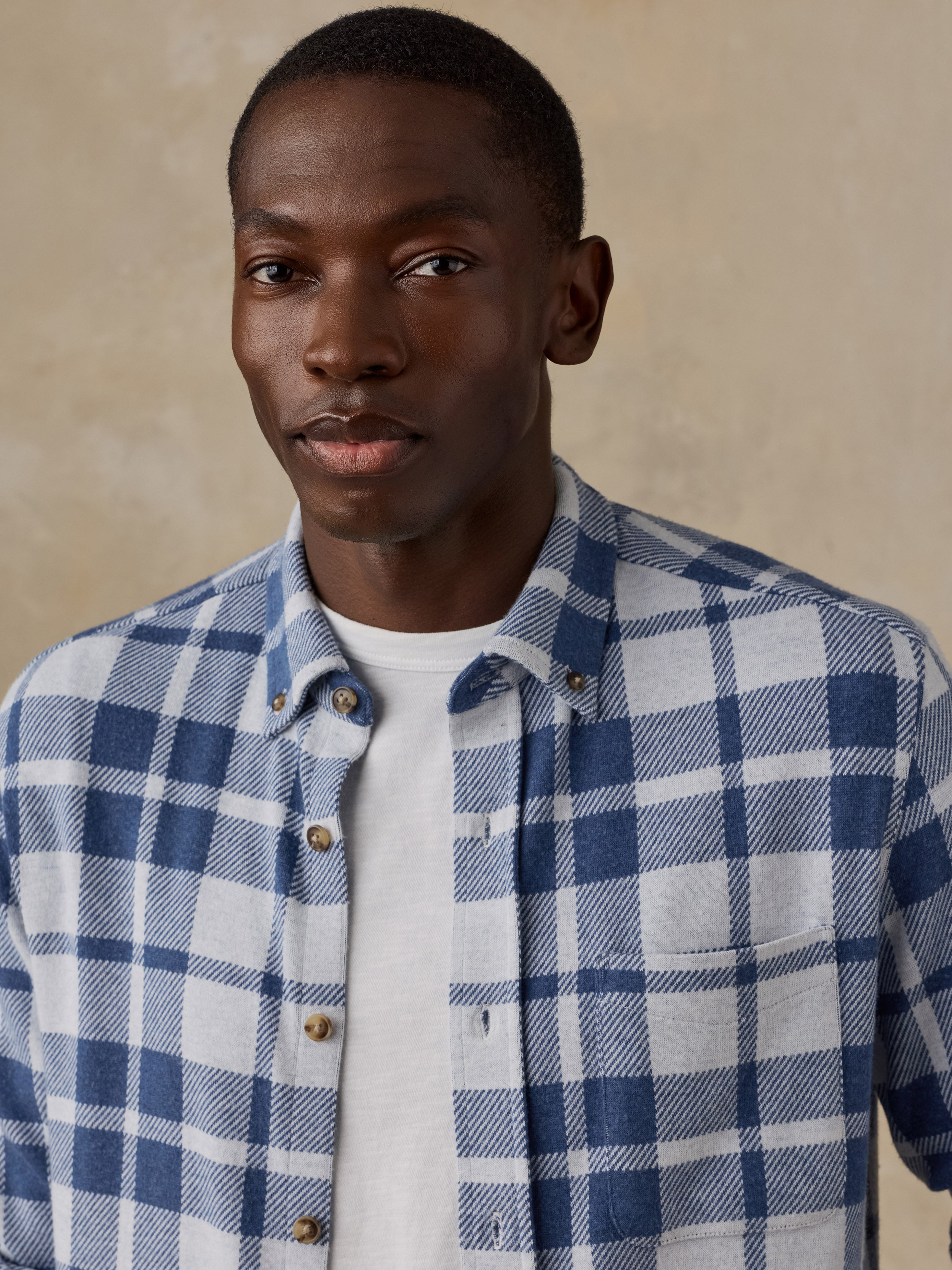 A man wearing a blue and white plaid button-down shirt over a white t-shirt, photographed against a neutral beige textured background.
