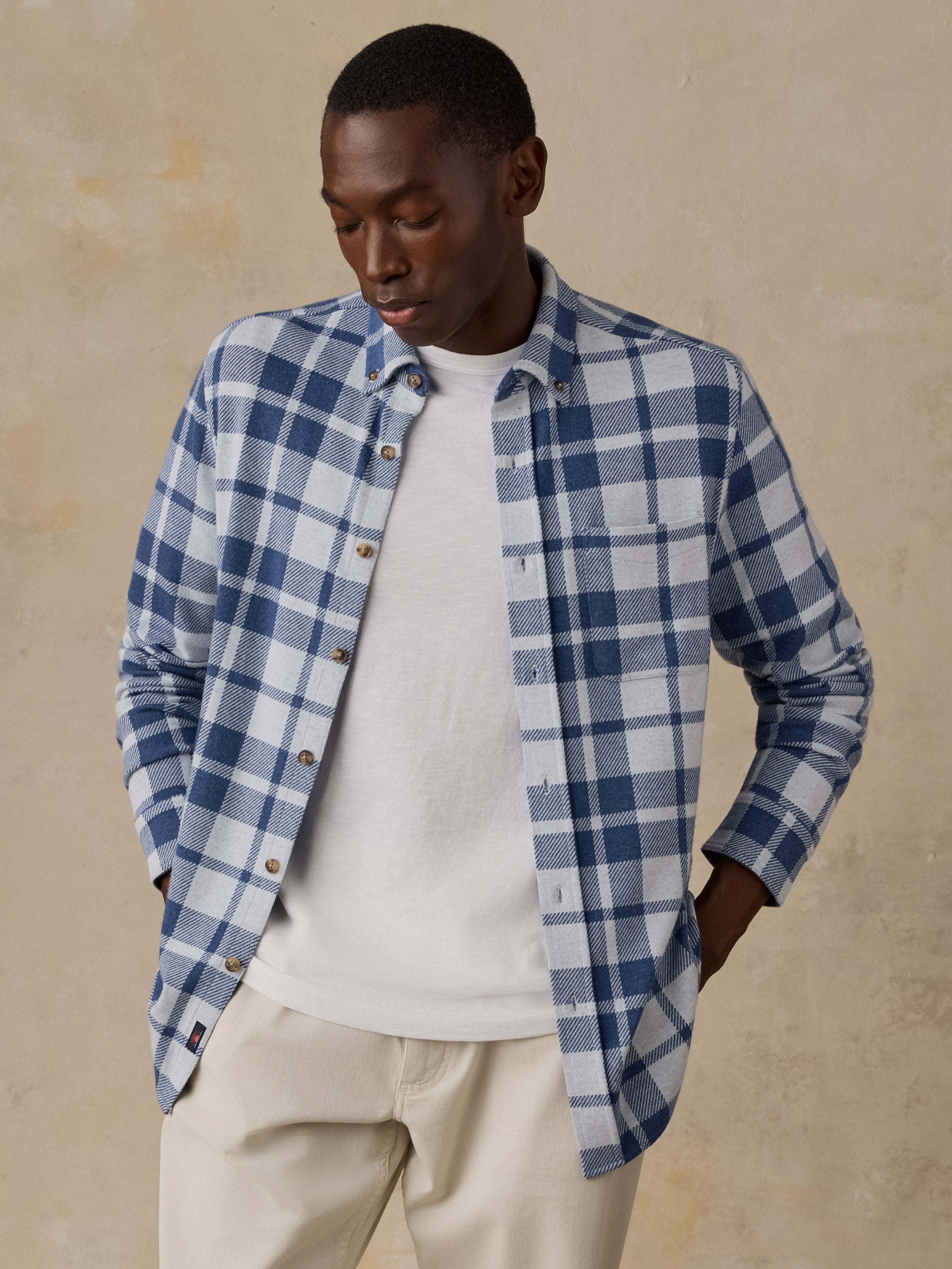 A man wearing a blue and white plaid button-down shirt over a white t-shirt and beige pants, standing against a textured beige wall.
