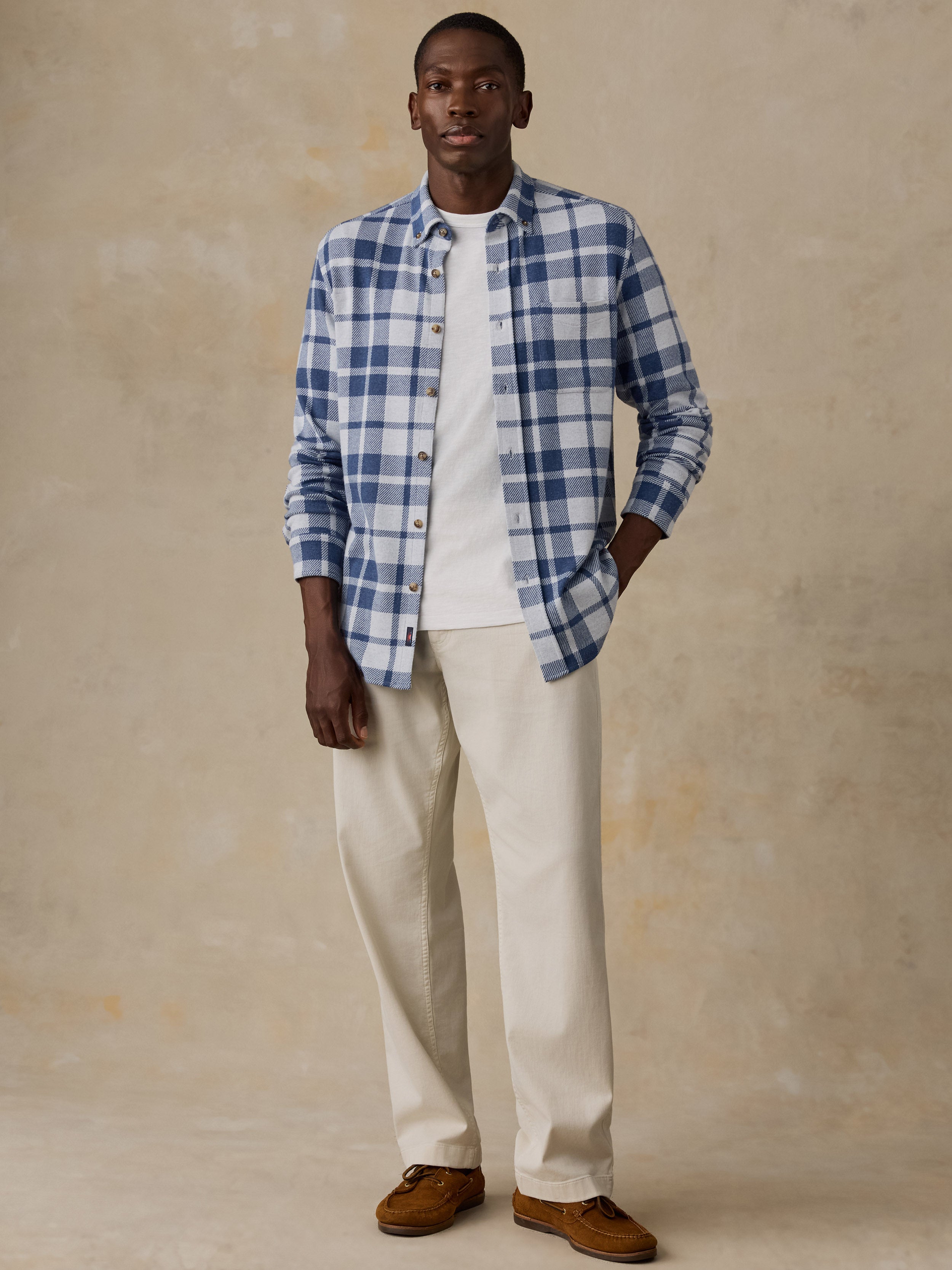 A man modeling a blue and white plaid button-down shirt over a white t-shirt, paired with cream trousers and brown suede shoes, against a textured beige studio backdrop.