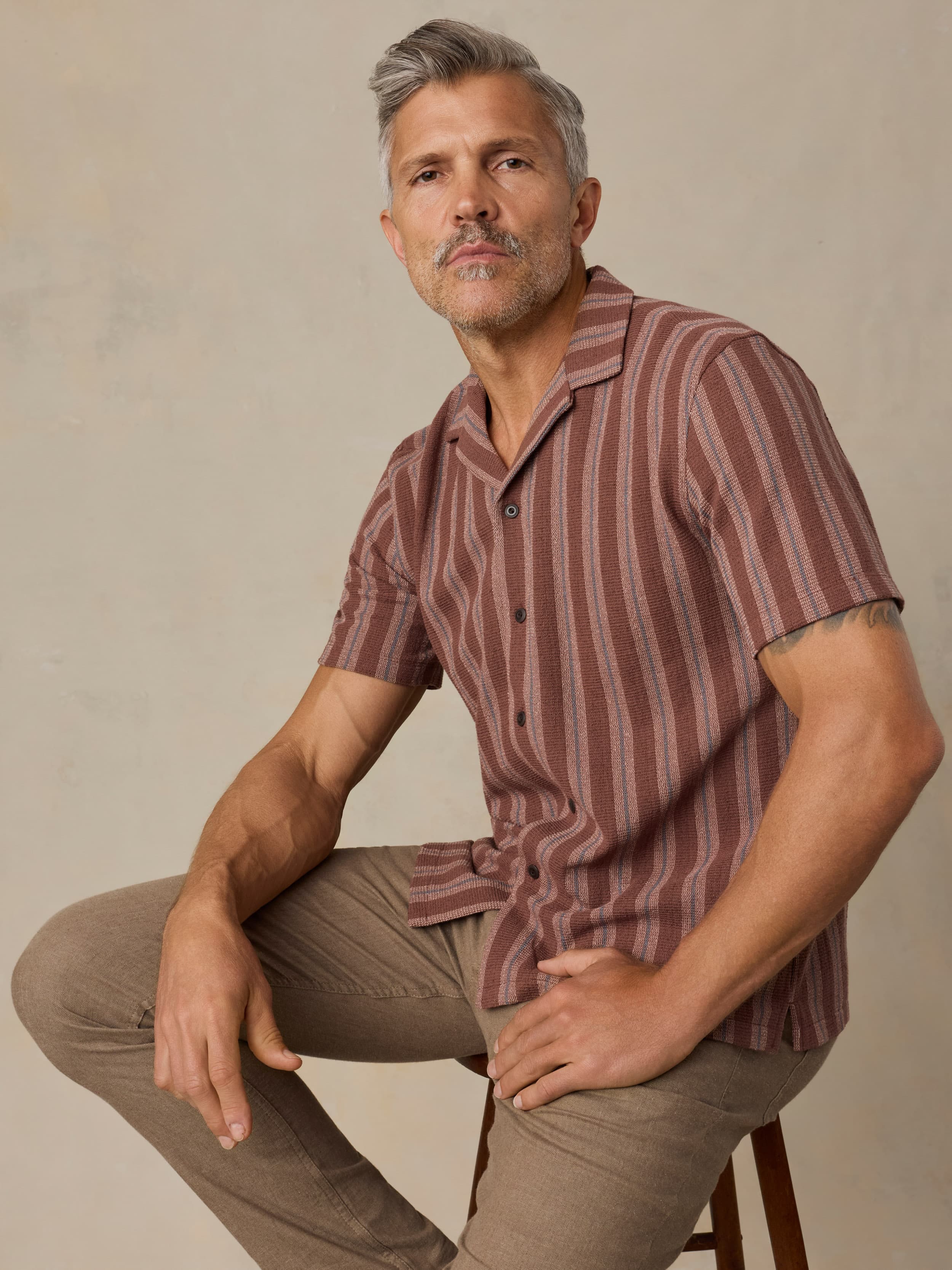 Man wearing a red striped shirt and brown pants sitting on a wooden stool against a beige background