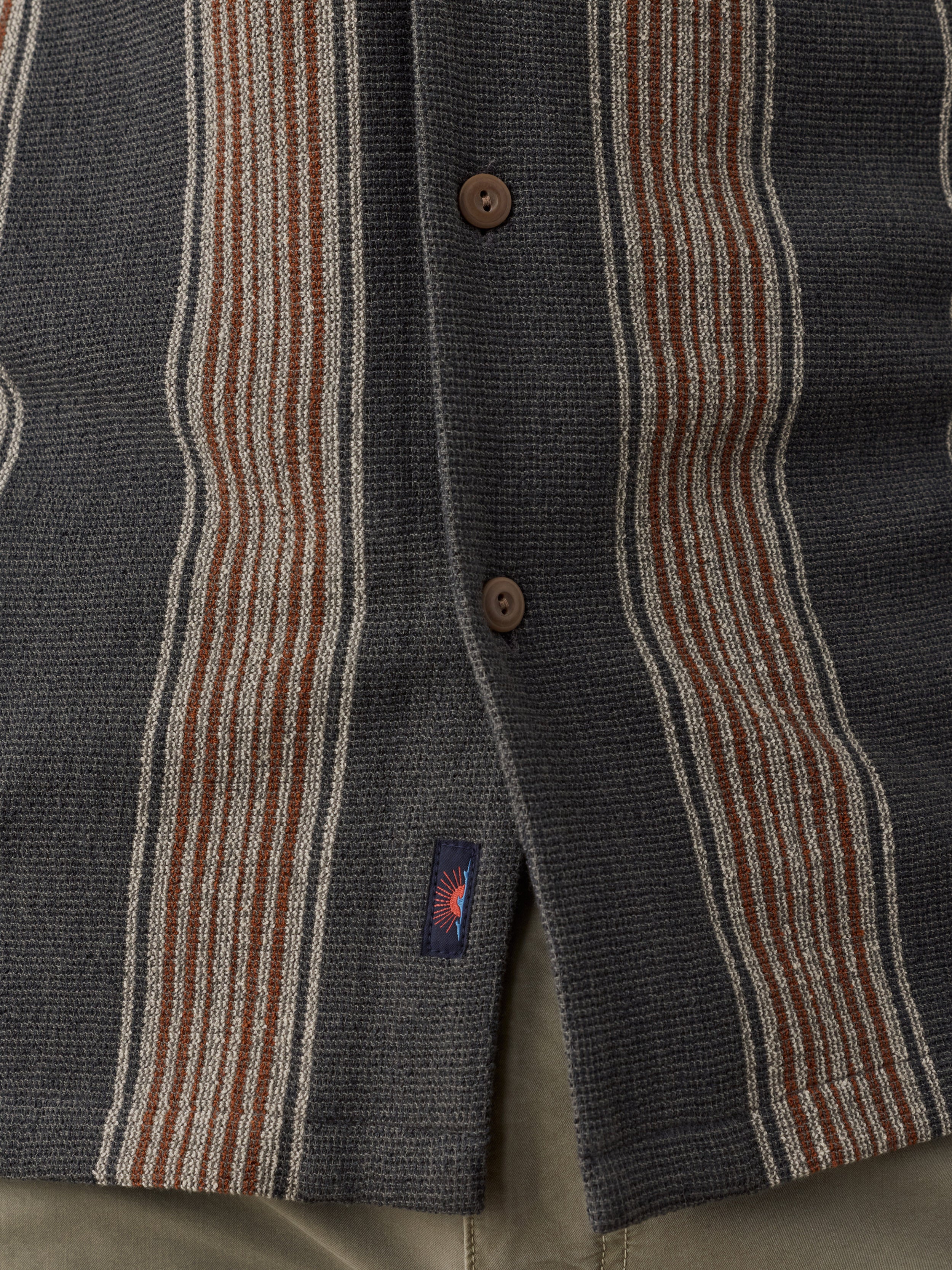 Close-up of a striped knit button-front garment, likely a cardigan or sweater, with vertical bands of dark gray, rust brown, cream, and muted red. A small woven Faherty brand tag near the hem.